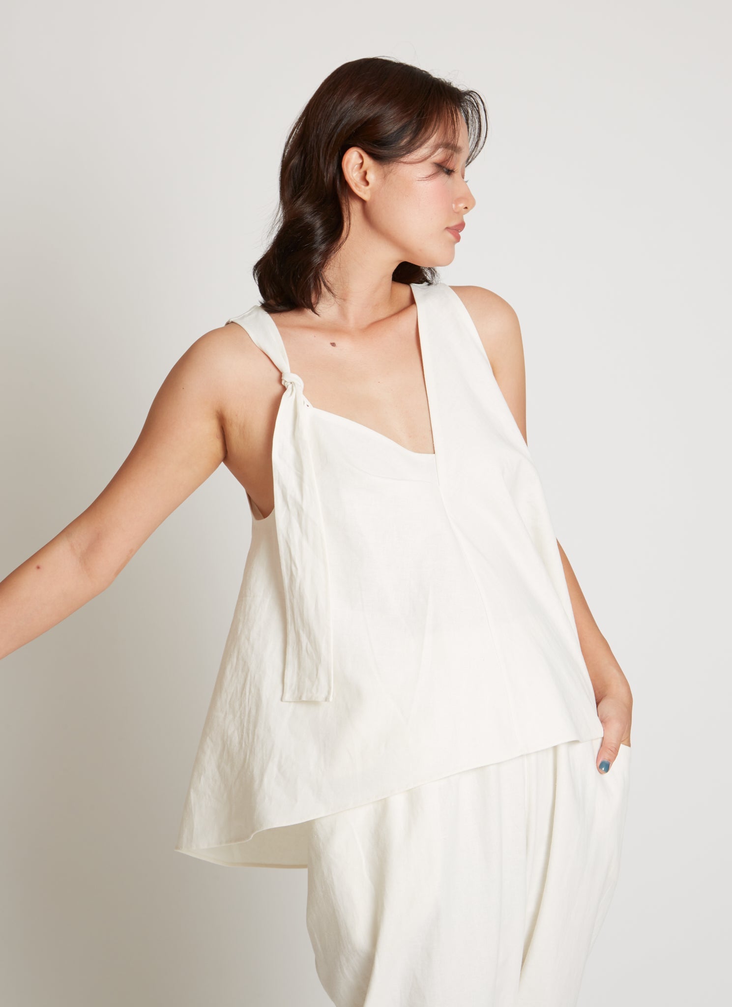 Front view of a woman standing wearing spring fashion in a Kaarem SS26 White Carrara Tie-Shoulder Asymmetrical Pleated Top at Alas Eius.