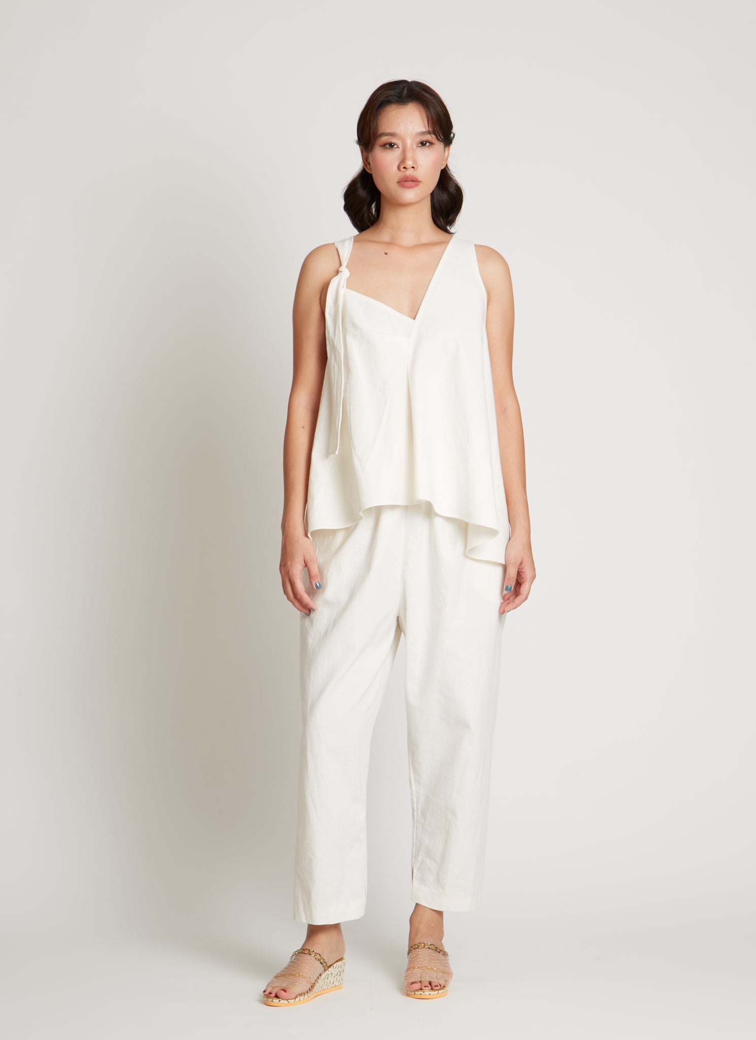 Full length front view of a woman standing wearing spring fashion in a Kaarem SS26 White Carrara Tie-Shoulder Pleated Top at Alas Eius.