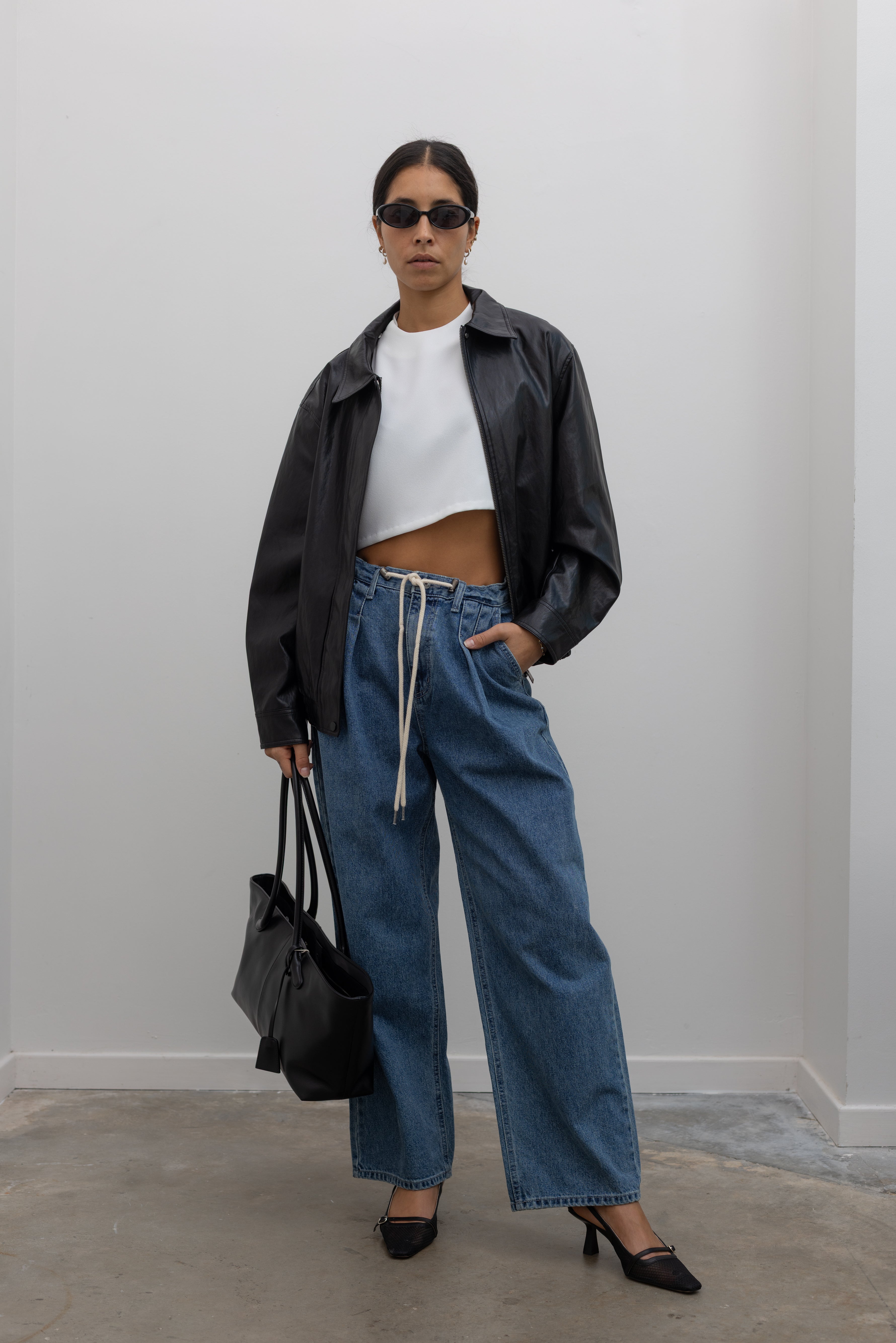 Close-up front view of a woman wearing capsule wardrobe essential style in a Kaarem White Raised Collar Wind Chime Cropped Top paired with light wash denim drawstring waist jeans and black zip jacket at Alas Eius.