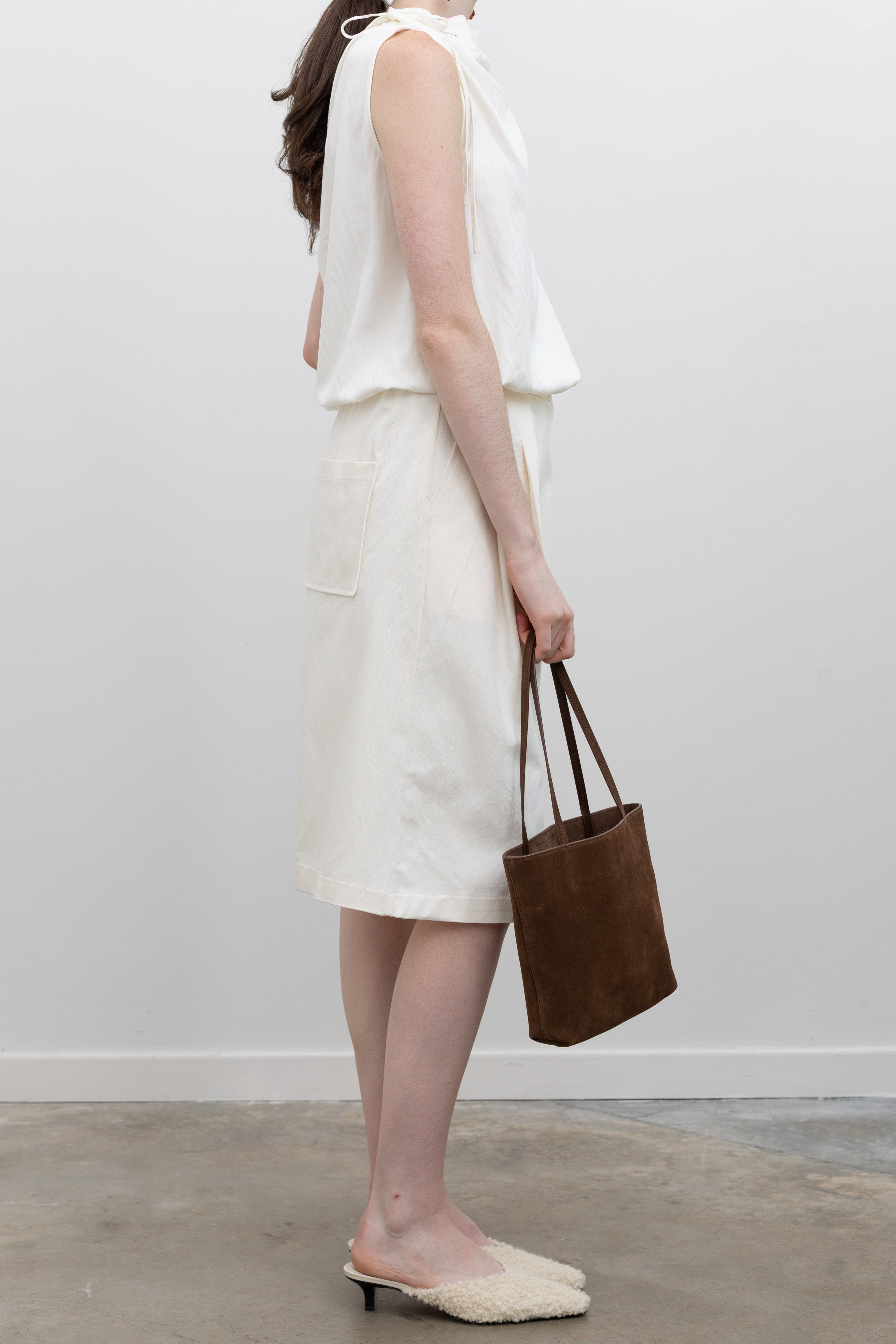 Woman wearing white bermuda shorts carrying a small tote by Kallmeyer in rich brown suede, dual flat shoulder straps, and a permanent canvas drawstring pouch opening with leather toggles at Alas Eius. Made in Italy. 