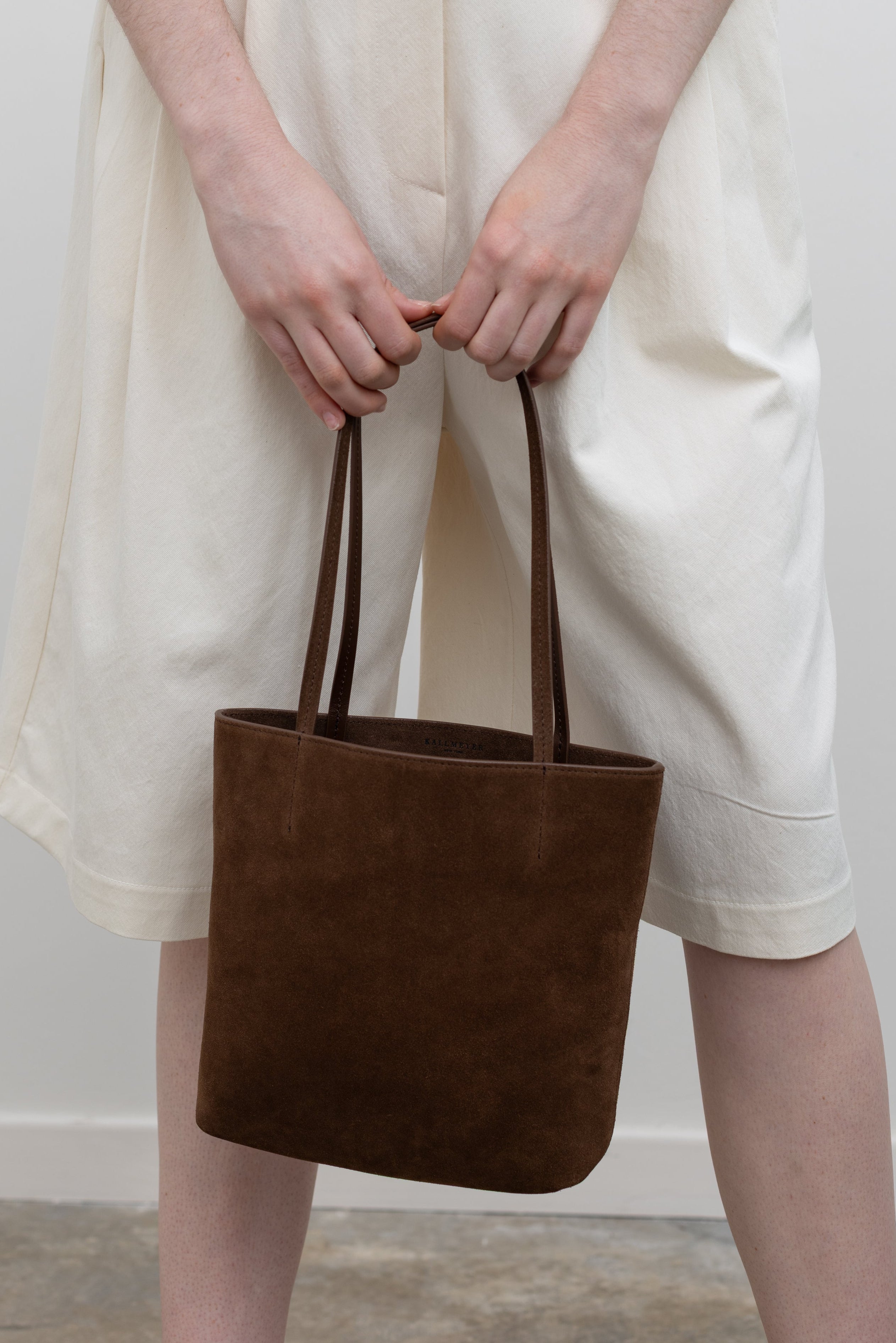 Woman wearing white bermuda shorts carrying a small tote by Kallmeyer in rich brown suede, dual flat shoulder straps, and a permanent canvas drawstring pouch opening with leather toggles at Alas Eius. Made in Italy. 