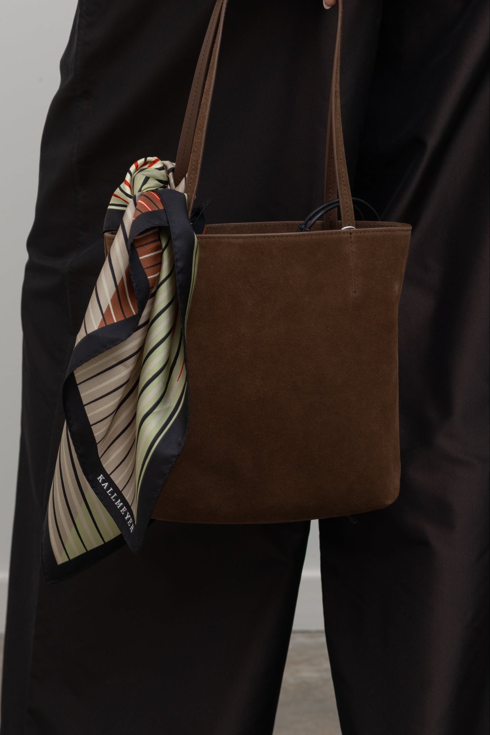 A classic small square Kallmeyer silk twill scarf featuring a gradient stripe with tonal red, grey, green, cream colors  tied on a Kallmeyer chocolate brown suede brunch tote bag at Alas Eius. 