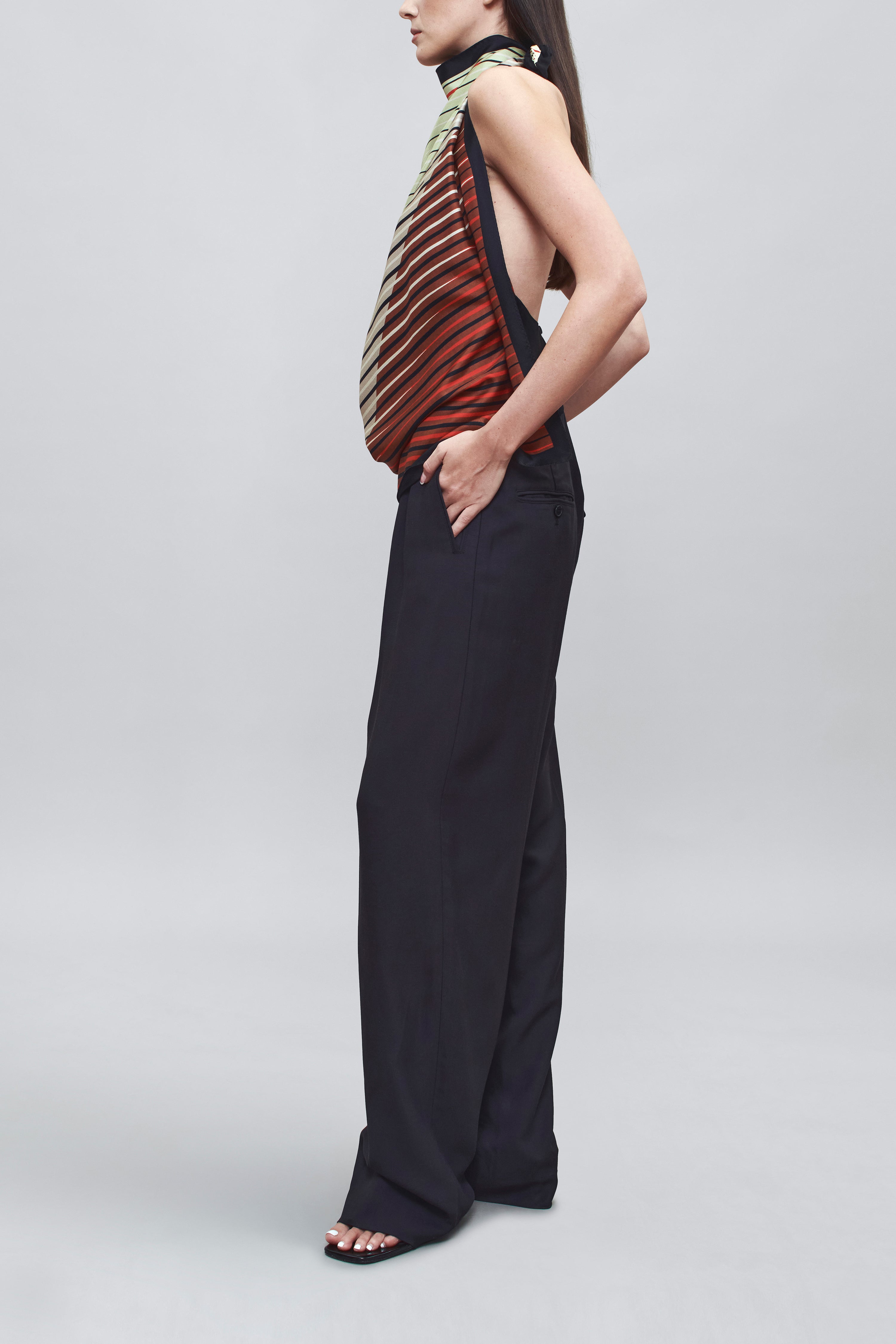 Side view of a woman wearing chic spring summer fashion in a colorful Kallmeyer Neutral Silk Twill Fluted Scarf Top featuring a gradient stripe print, soft cowl neckline, and a deep open back with fitted panel at lower back paired with navy blue trousers at an Alas Eius store.