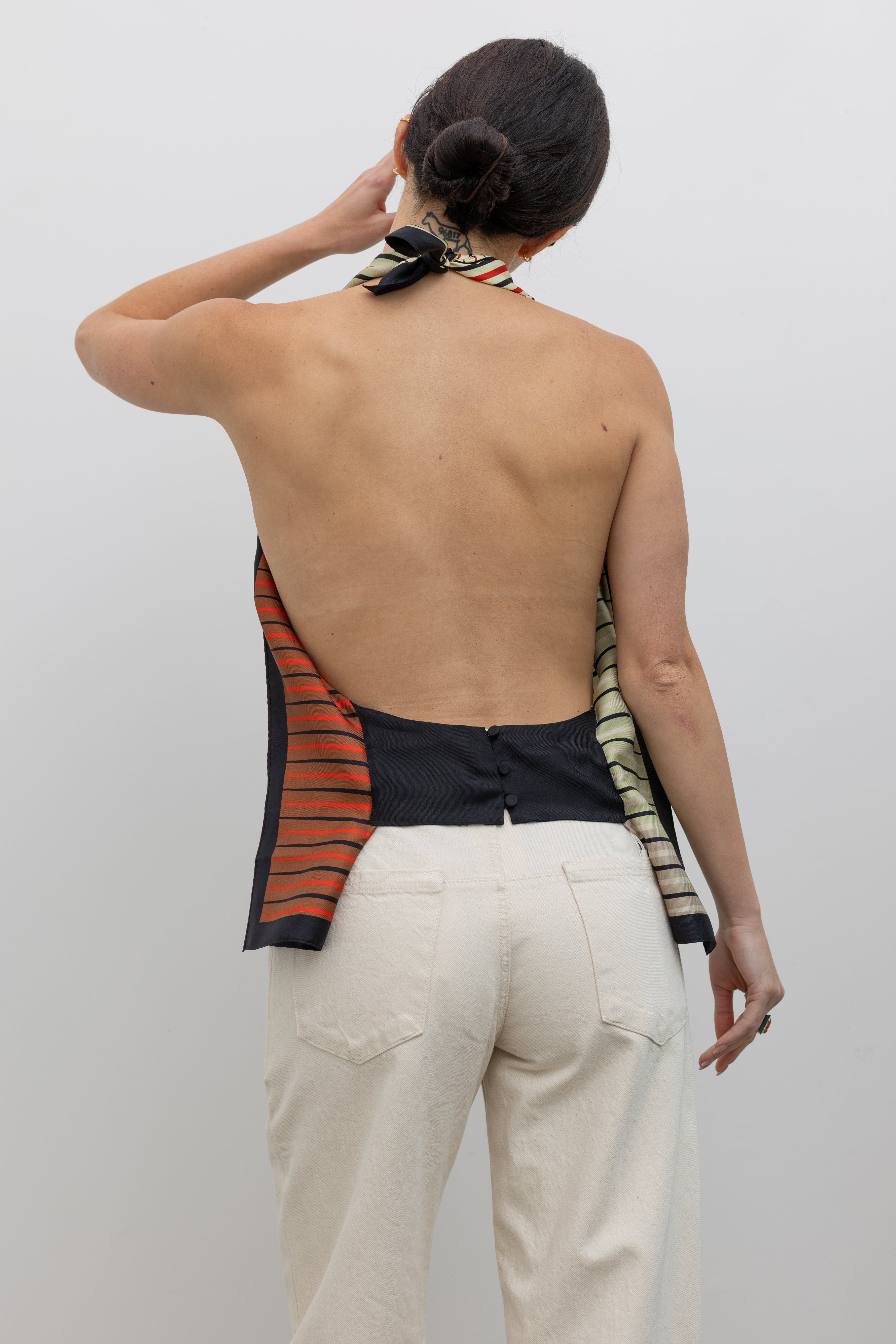 Close-up back view of a woman wearing the perfect summer vacation wardrobe staple in a colorful Kallmeyer Neutral Silk Twill Fluted Scarf Top featuring a gradient stripe print, soft cowl neckline, and a deep open back with fitted panel at lower back paired with ivory colored denim jeans and strappy black sandals at an Alas Eius store.