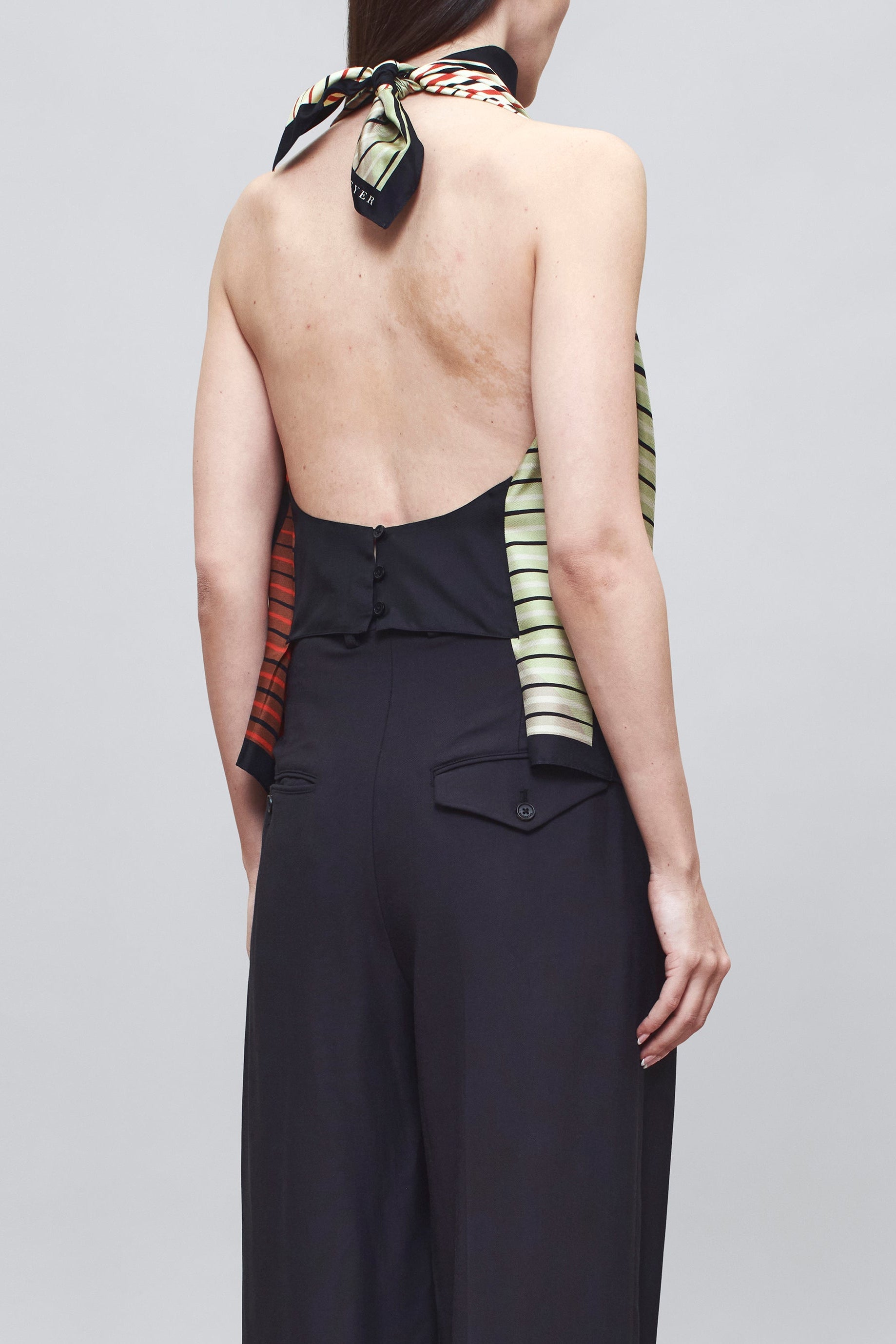 Back view of a woman wearing chic spring summer fashion in a colorful Kallmeyer Neutral Silk Twill Fluted Scarf Top featuring a gradient stripe print, soft cowl neckline, and a deep open back with fitted panel at lower back paired with navy blue trousers at an Alas Eius store.