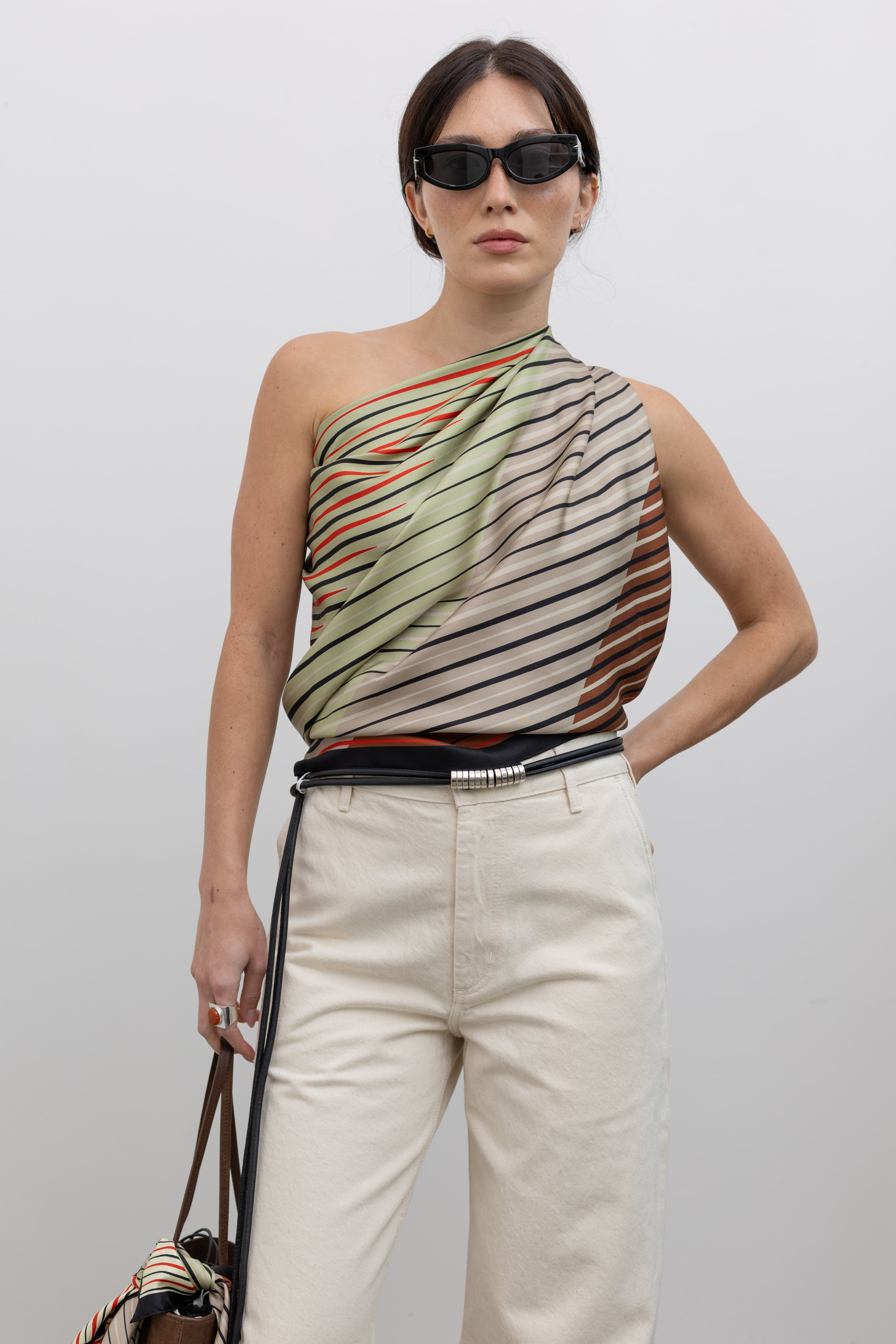 Close-up front view of a woman wearing modern minimalist spring summer fashion in a colorful Kallmeyer Neutral Silk Twill Fluted Scarf Top featuring a gradient stripe print, soft cowl neckline, and a deep open back with fitted panel at lower back paired with ivory colored denim jeans and strappy black sandals at an Alas Eius store.