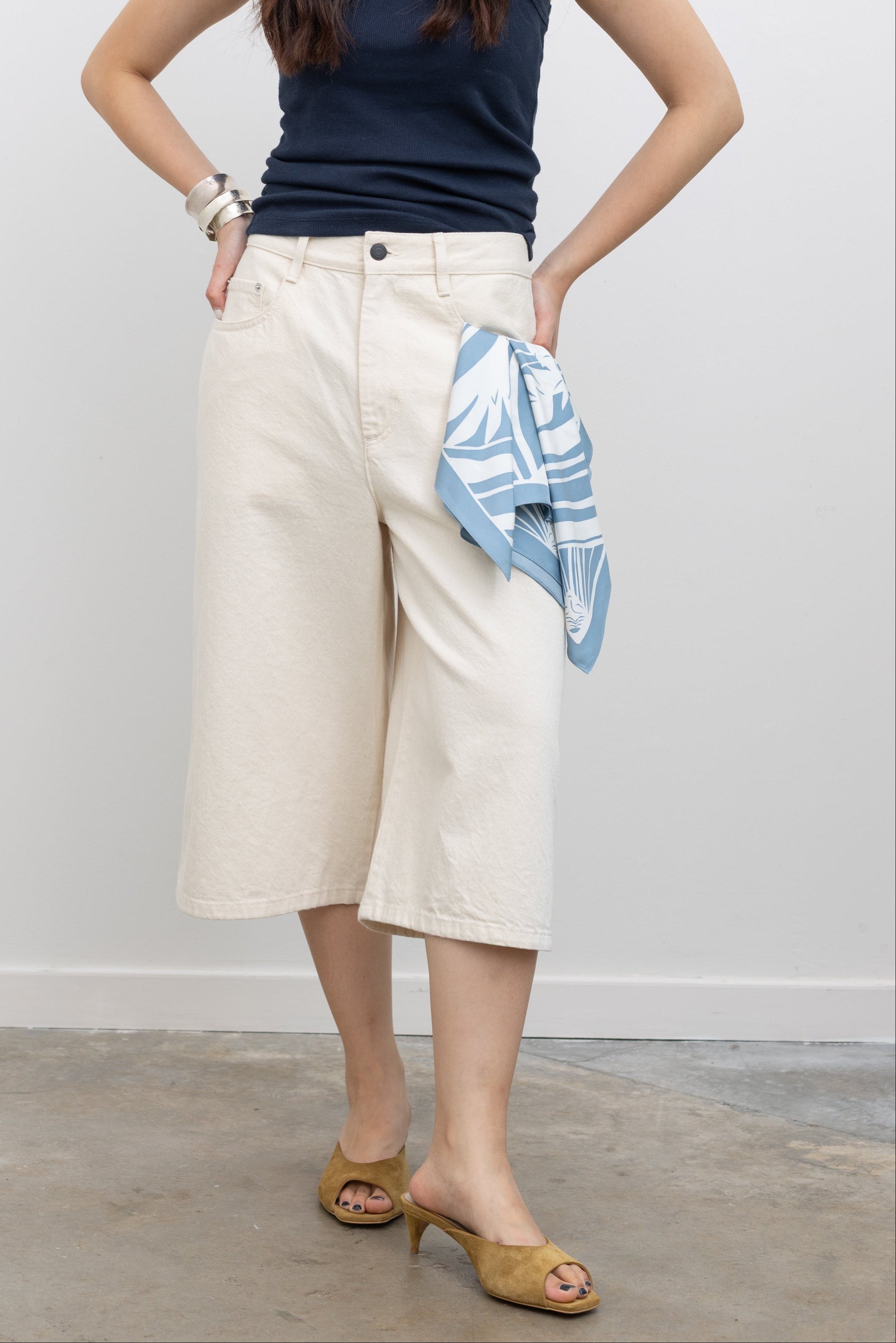 Model wearing chic Kallmeyer ecru hayes shorts with a wide-leg, high-rise waist, and cropped length styled with a navy sleeveless top, tan heeled sandals, and a blue printed scarf tucked into the pocket at Alas Eius.