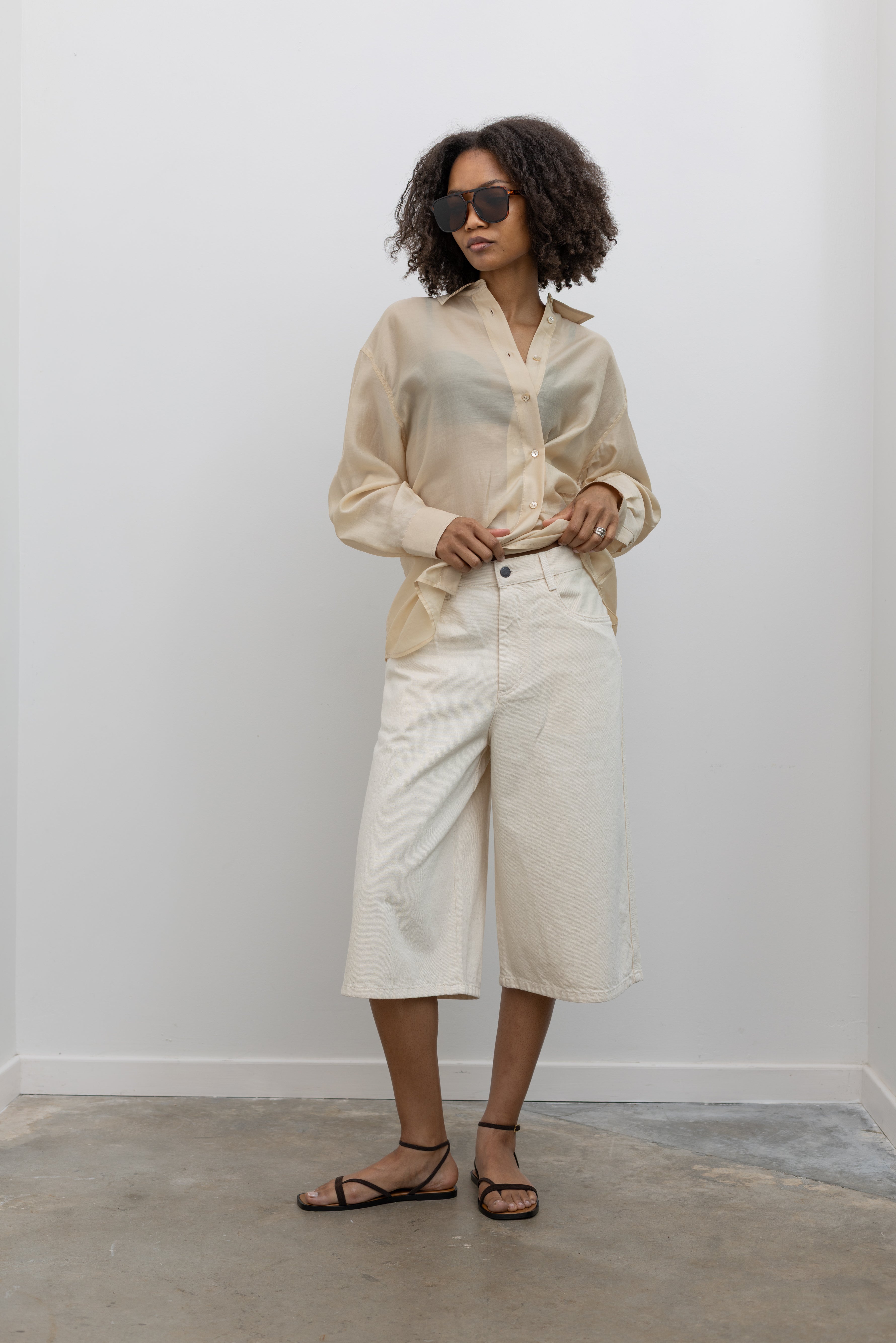 Full length front view of a woman standing wearing elevated women's spring summer fashion a pair of Kallmeyer ecru cotton Hayes wide-leg denim shorts with a structured leg that stops just below the knee paired with a Mijeong Park semi sheer beige button down shirt and strappy sandals at an Alas Eius store. Made in Portugal.