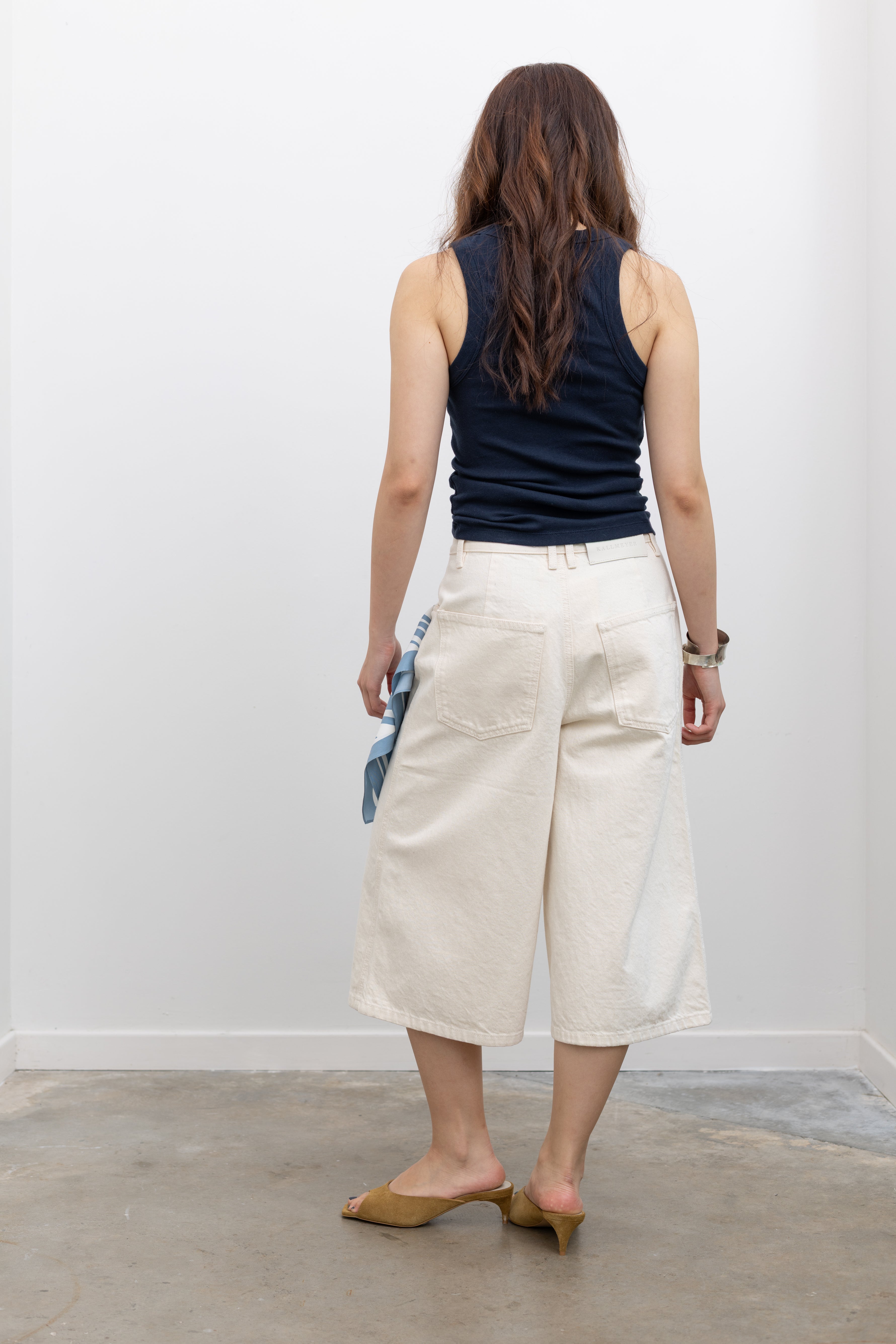 Model wearing Kallmeyer chic high-waisted cream cotton cropped denim trousers with a relaxed wide-leg fit, paired with a navy sleeveless top and tan suede heeled sandals at Alas Eius. 