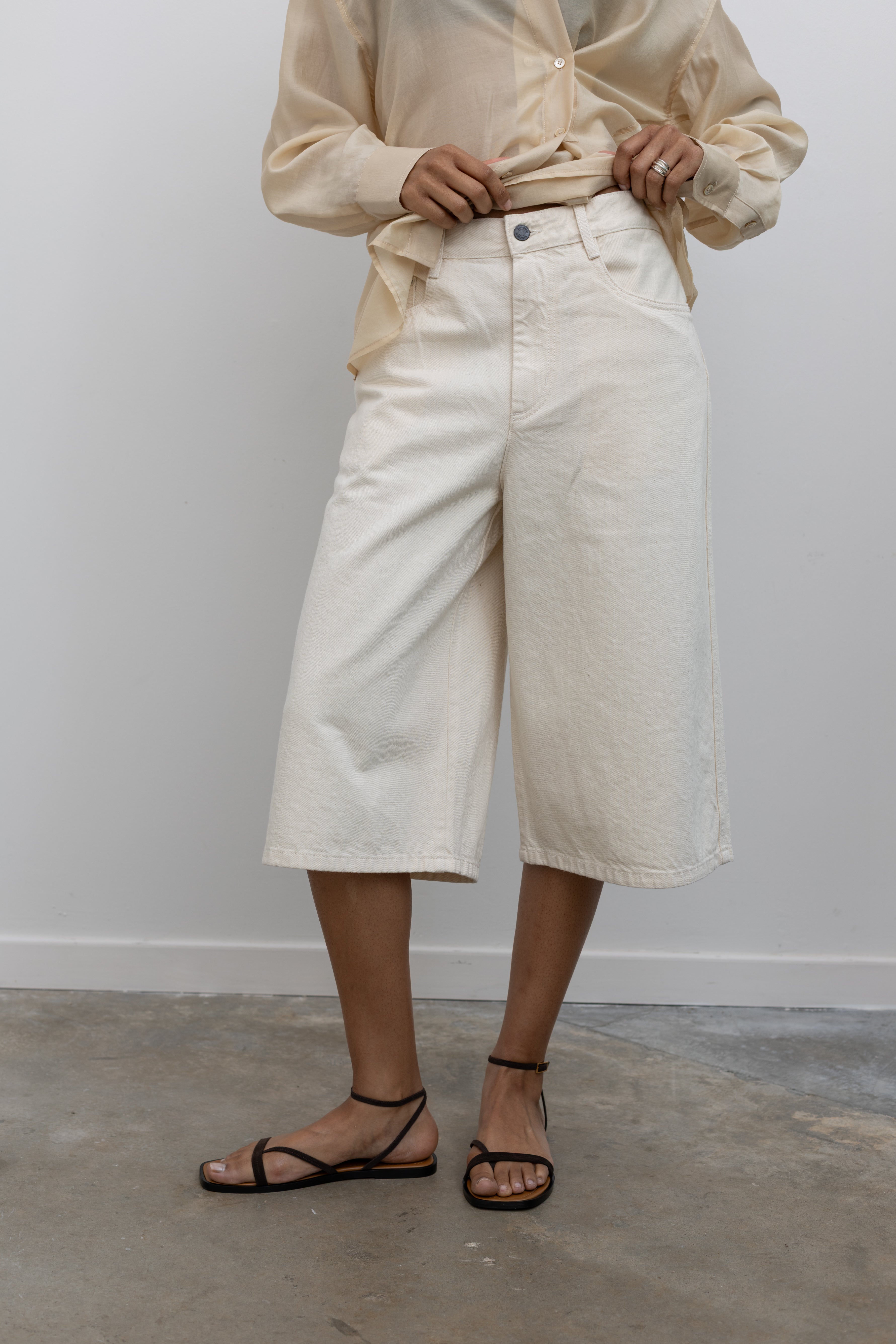 Close-up front view of a woman standing wearing elevated women's spring summer fashion a pair of Kallmeyer ecru cotton Hayes wide-leg denim shorts with a structured leg that stops just below the knee paired with strappy sandals at an Alas Eius store. Made in Portugal.