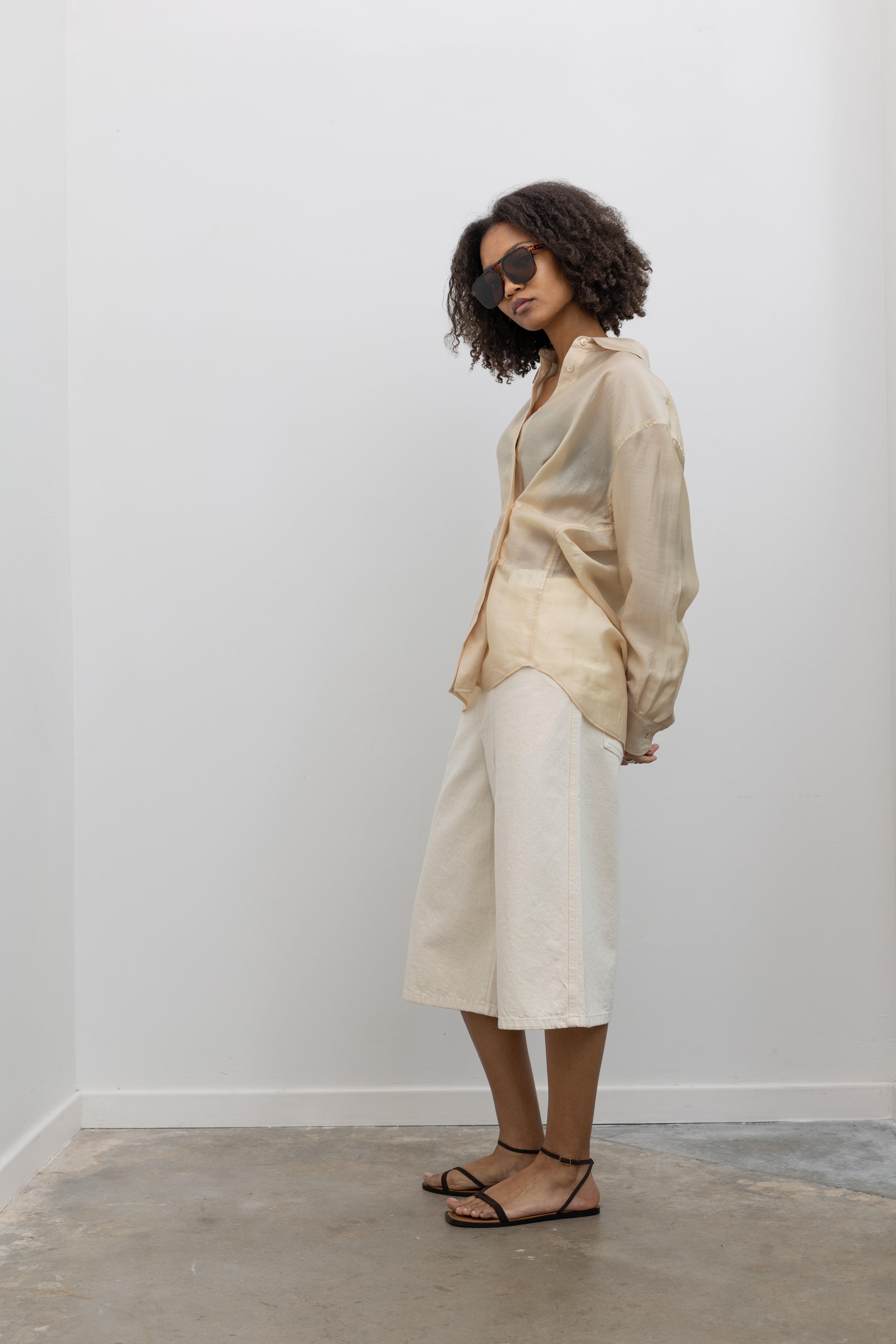 Full length 3/4 view of a woman standing wearing elevated and refined casual spring summer fashion a pair of Kallmeyer ecru cotton Hayes wide-leg denim shorts with a structured leg that stops just below the knee paired with a Mijeong Park semi sheer beige button down shirt and strappy sandals at an Alas Eius store. Made in Portugal.