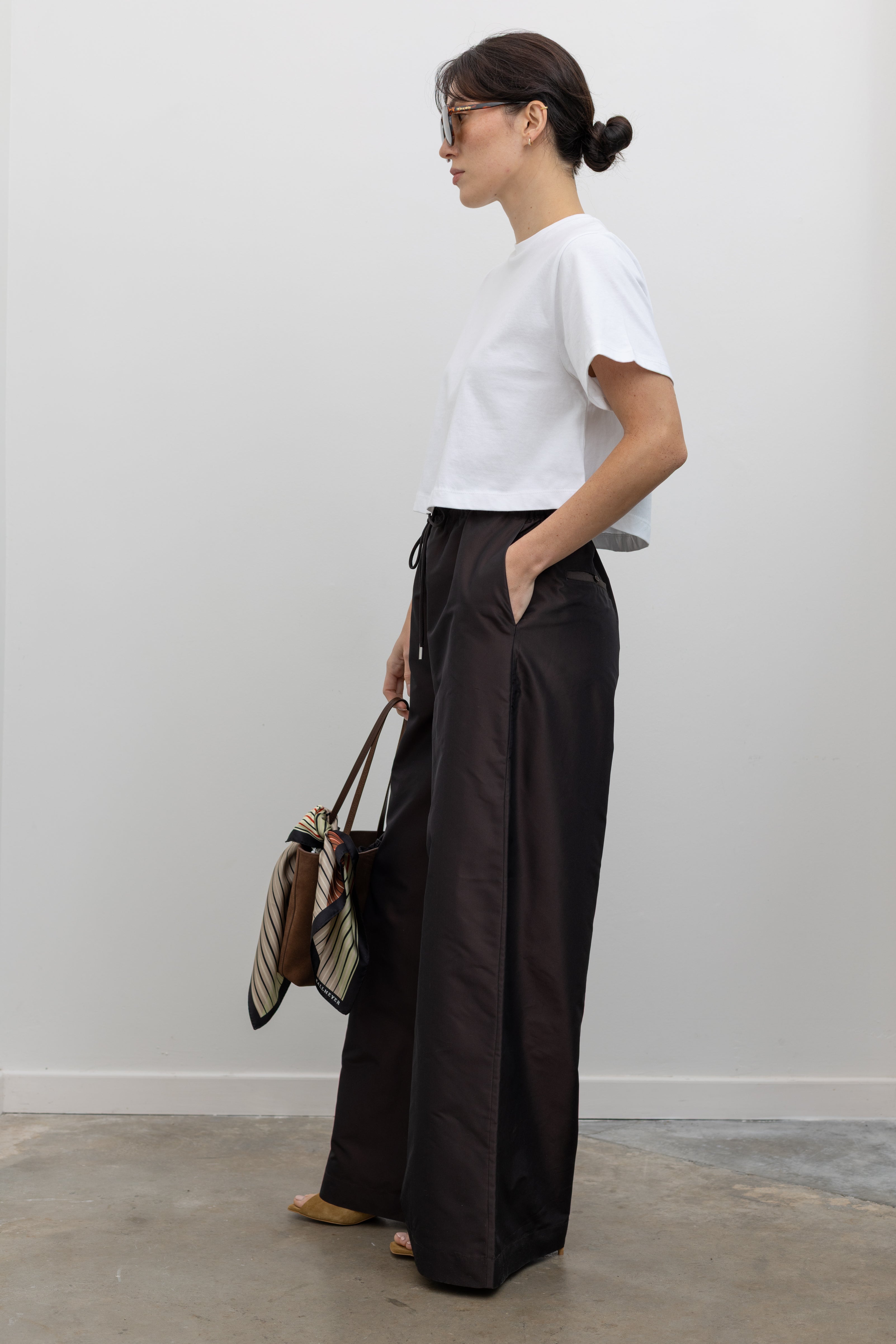 Full length side view of a woman wearing versatile and contemporary fashion in Kallmeyer Espresso Silk Taffeta Wesley Drawstring Pants featuring a hardware drawstring waist and structured wide legs at an Alas Eius store. Made in New York.
