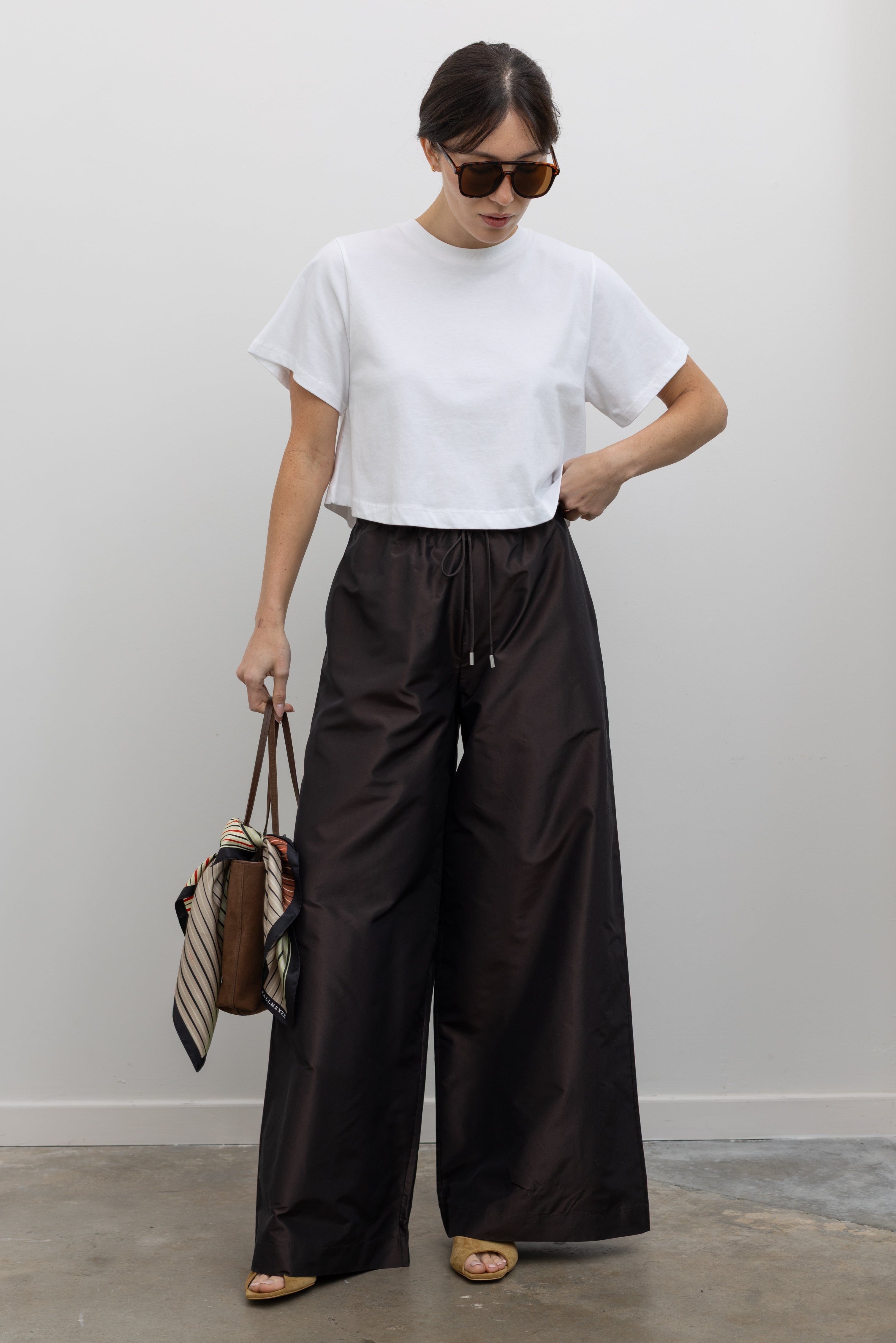 Full length front view of a woman wearing chic and contemporary fashion in Kallmeyer Espresso Silk Taffeta Wesley Drawstring Pants featuring a hardware drawstring waist and structured wide legs paired with an Alas Eius Label white cropped  t-shirt at an Alas Eius store. Made in New York.