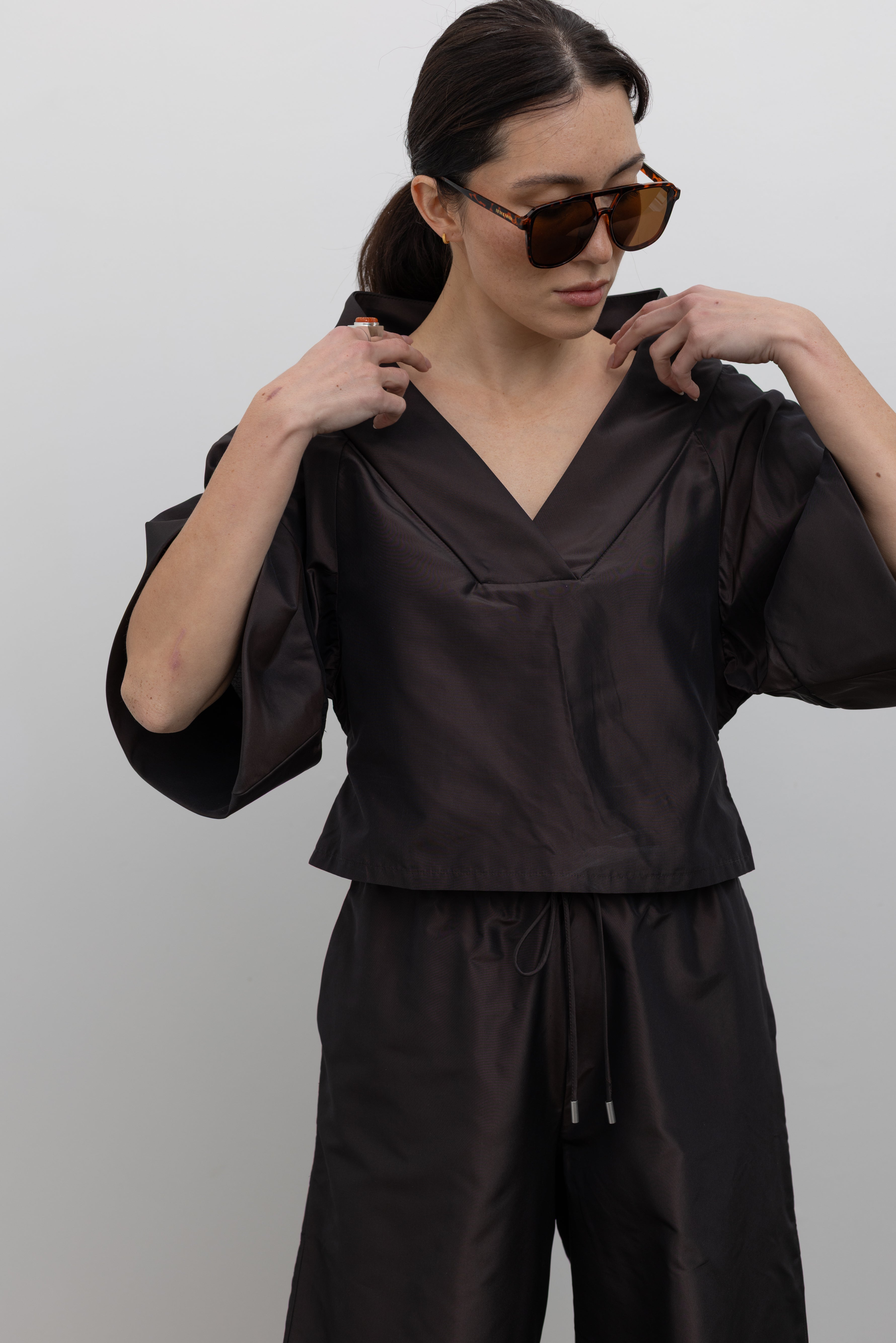 Close-up front view of a woman wearing chic and modern fashion in a Kallmeyer Espresso Silk Taffeta Winslow Top featuring a dramatic structured v-neckline and oversized cuffed tunic sleeves; paired with Espresso Silk Taffeta Wesley Drawstring Pant featuring a hardware drawstring waist and structured wide legs at an Alas Eius store. Made in New York.