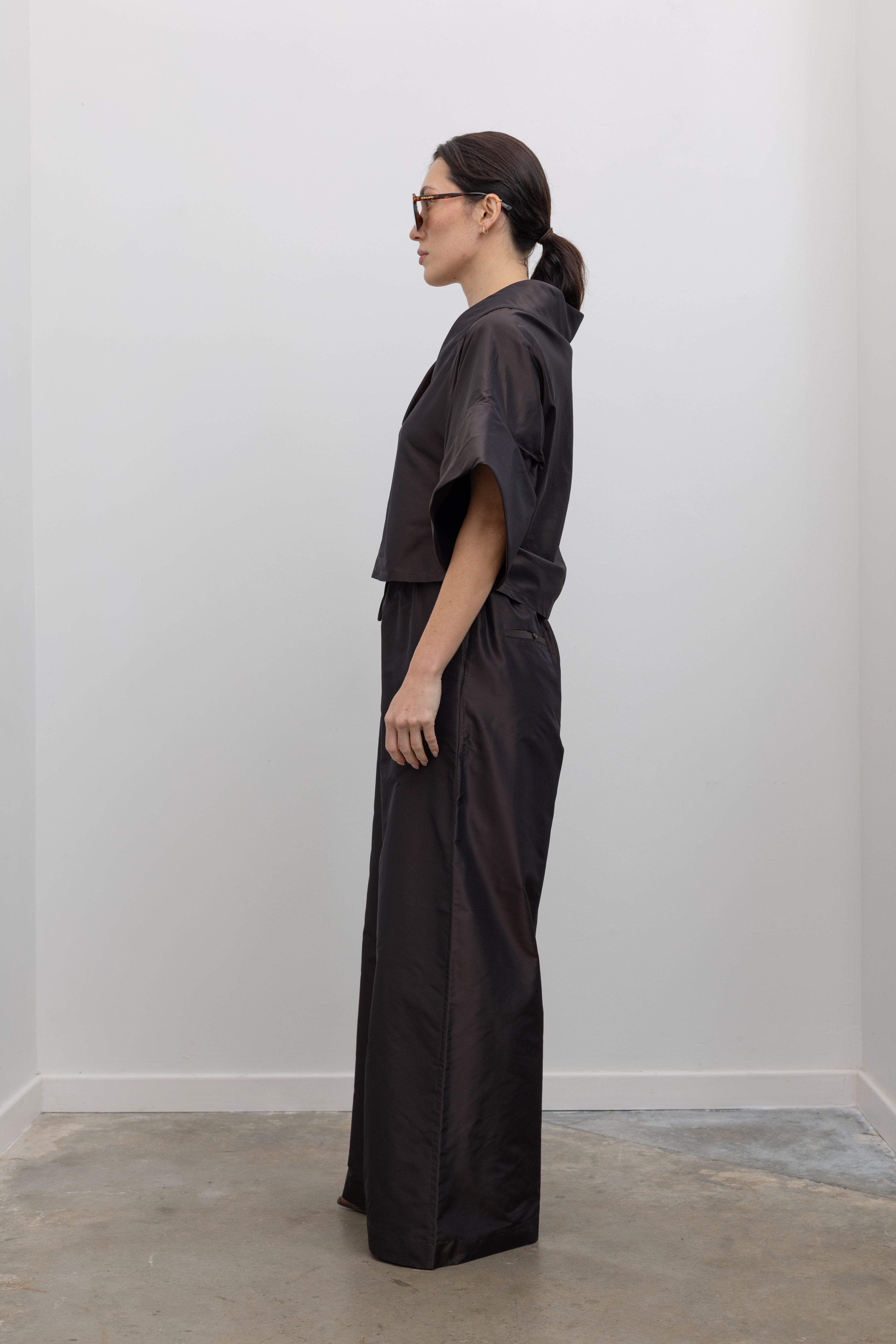 Full length side view of a woman wearing chic and contemporary fashion in a Kallmeyer Espresso Silk Taffeta Winslow Top featuring a dramatic structured v-neckline and oversized cuffed tunic sleeves; paired with Espresso Silk Taffeta Wesley Drawstring Pant featuring a hardware drawstring waist and structured wide legs at an Alas Eius store. Made in New York.