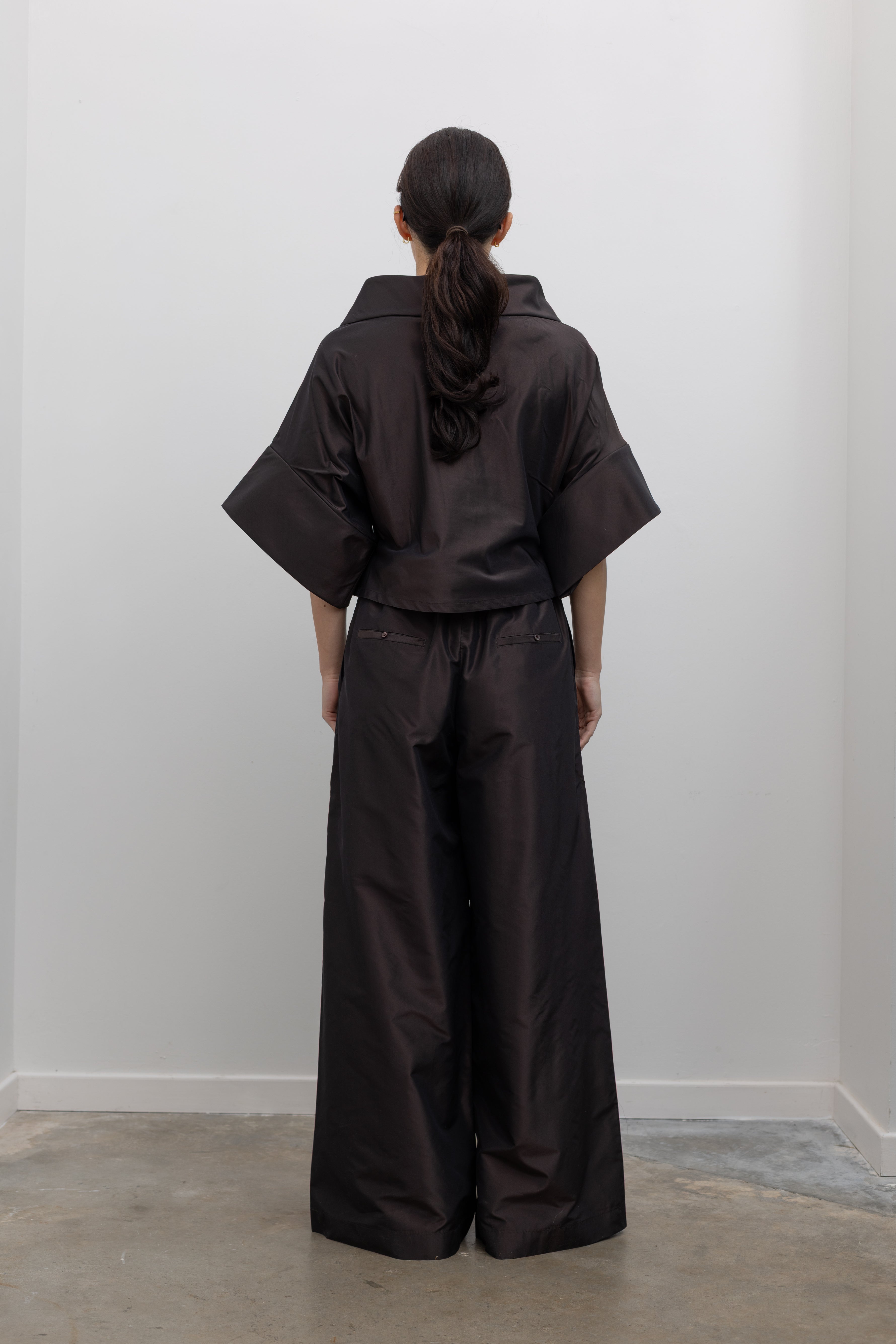 Full length back view of a woman wearing chic and contemporary fashion in a Kallmeyer Espresso Silk Taffeta Winslow Top featuring a dramatic structured v-neckline and oversized cuffed tunic sleeves; paired with Espresso Silk Taffeta Wesley Drawstring Pant featuring a hardware drawstring waist and structured wide legs at an Alas Eius store. Made in New York.