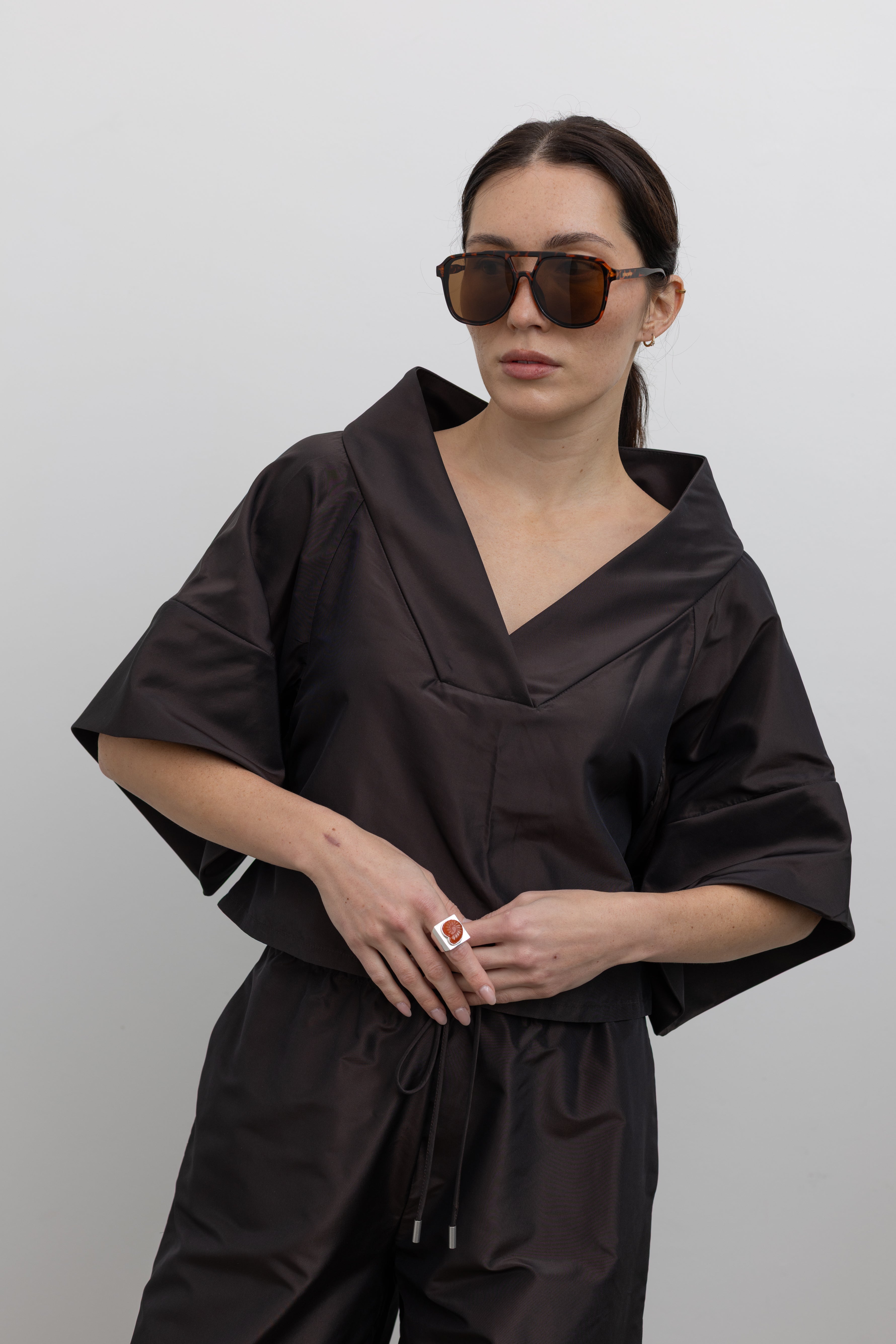 Front view of a woman wearing chic and contemporary fashion in a Kallmeyer Espresso Silk Taffeta Winslow Top featuring a crisp silk taffeta with dramatic structured v-neck and oversized cuffed tunic sleeves at an Alas Eius store. Made in New York.