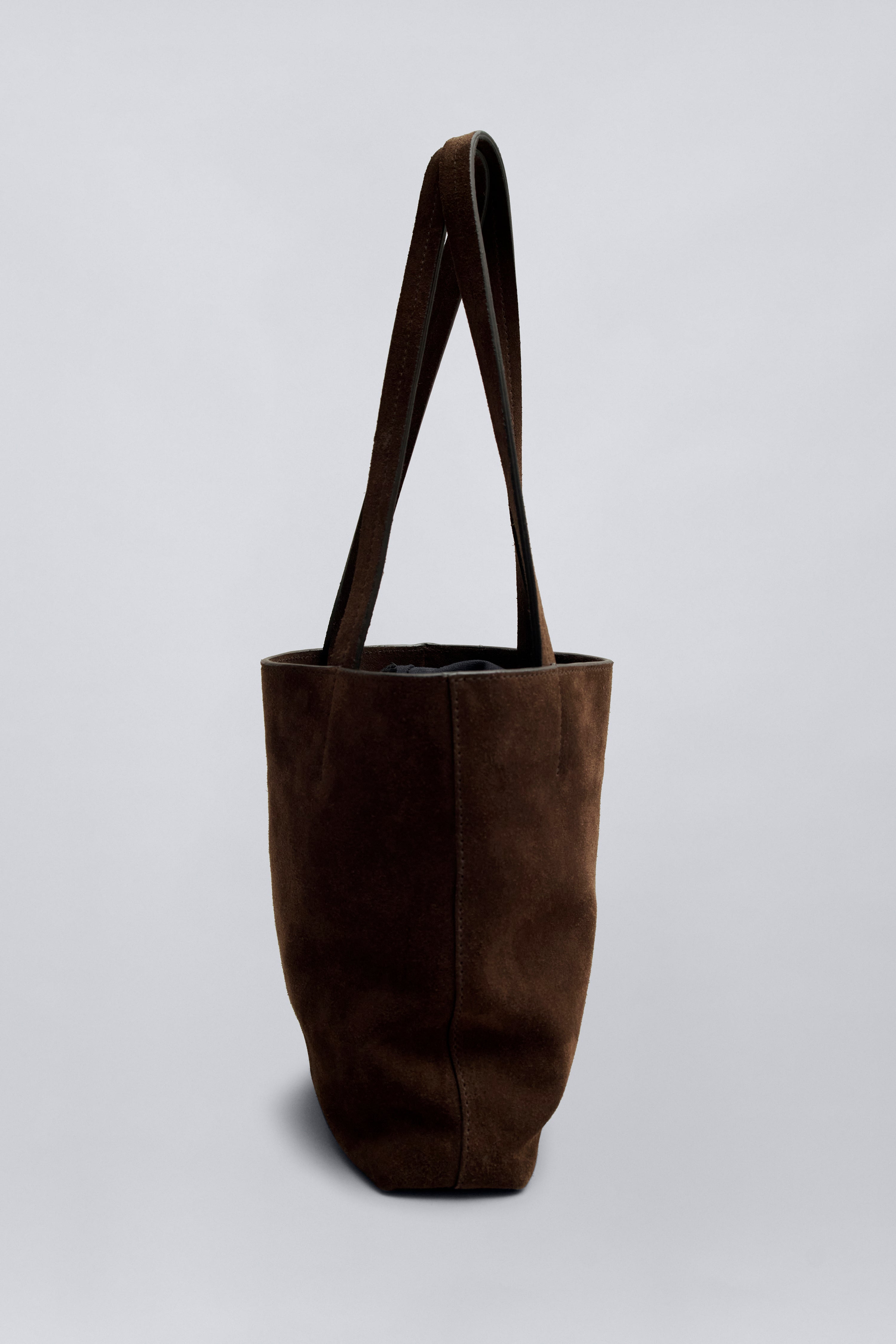 Side product view of a chic and elevated classic structured tote by Kallmeyer in rich brown suede, dual flat shoulder straps, and a permanent canvas drawstring pouch opening with leather toggles at Alas Eius. Made in Italy. 