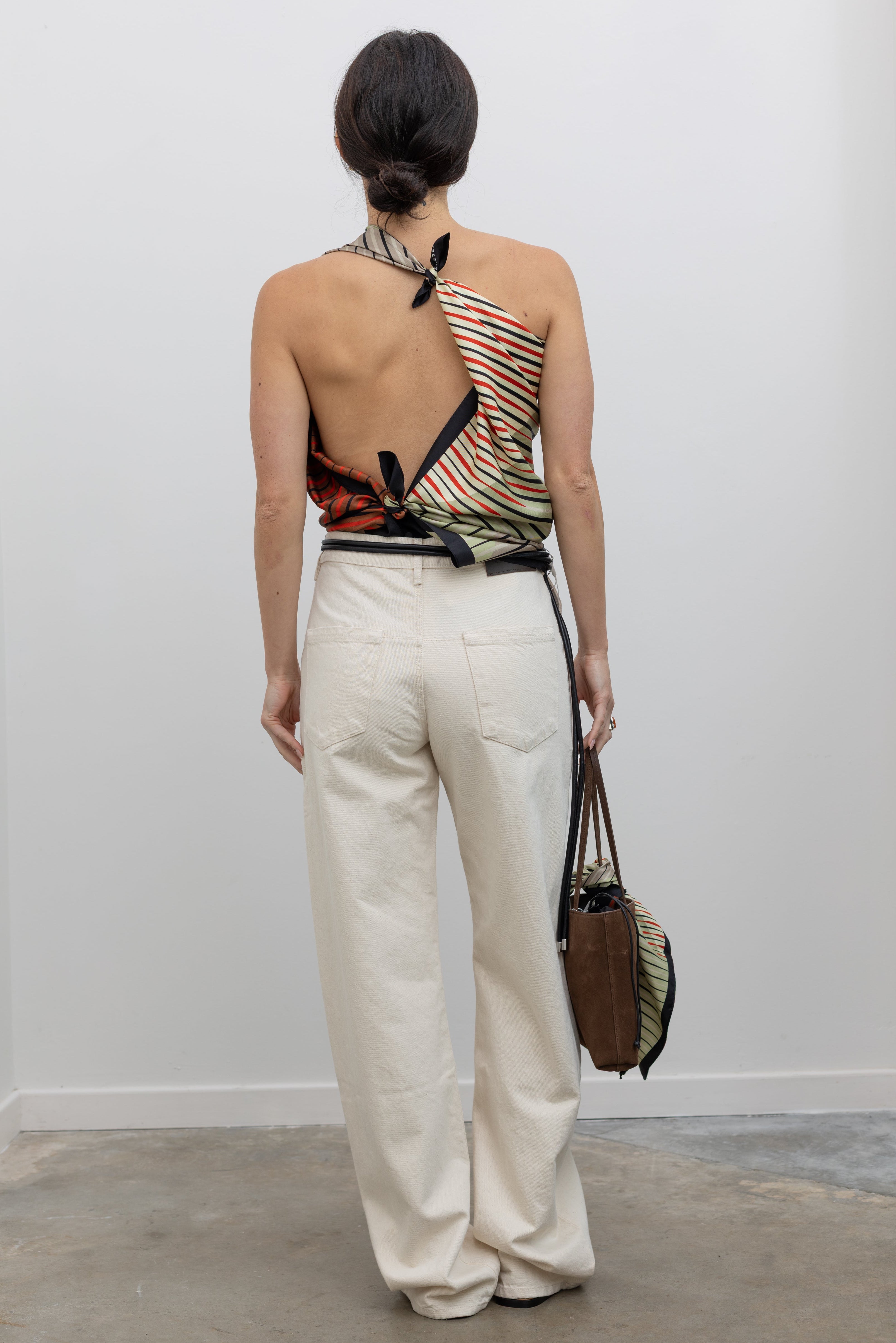 Full length back view of a woman wearing modern minimalist spring summer fashion in a Kallmeyer Neutral Silk Twill Fluted Scarf Top featuring a gradient stripe print, soft cowl neckline, and a deep open back with fitted panel at lower back paired with Kallmeyer Ecru Column Wide Leg Denim Jeans featuring a Mid-high rise, relaxed hip and thigh at an Alas Eius store. Made in Portugal. Made in New York, USA.