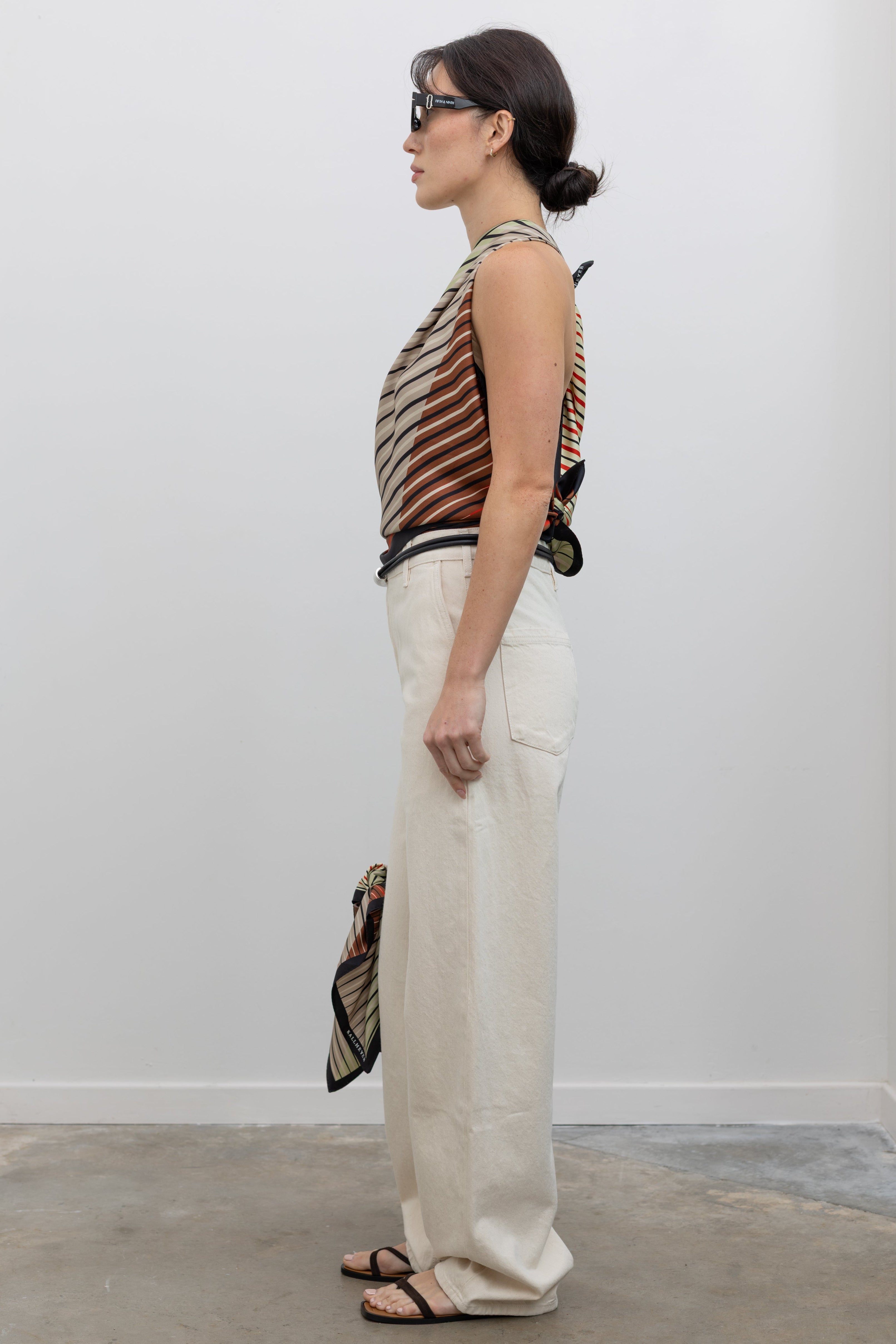 Full length side view of a woman wearing modern minimalist spring summer fashion in a Kallmeyer Neutral Silk Twill Fluted Scarf Top featuring a gradient stripe print, soft cowl neckline, and a deep open back with fitted panel at lower back paired with Kallmeyer Ecru Column Wide Leg Denim Jeans featuring a Mid-high rise, relaxed hip and thigh at an Alas Eius store. Made in Portugal. Made in New York, USA.