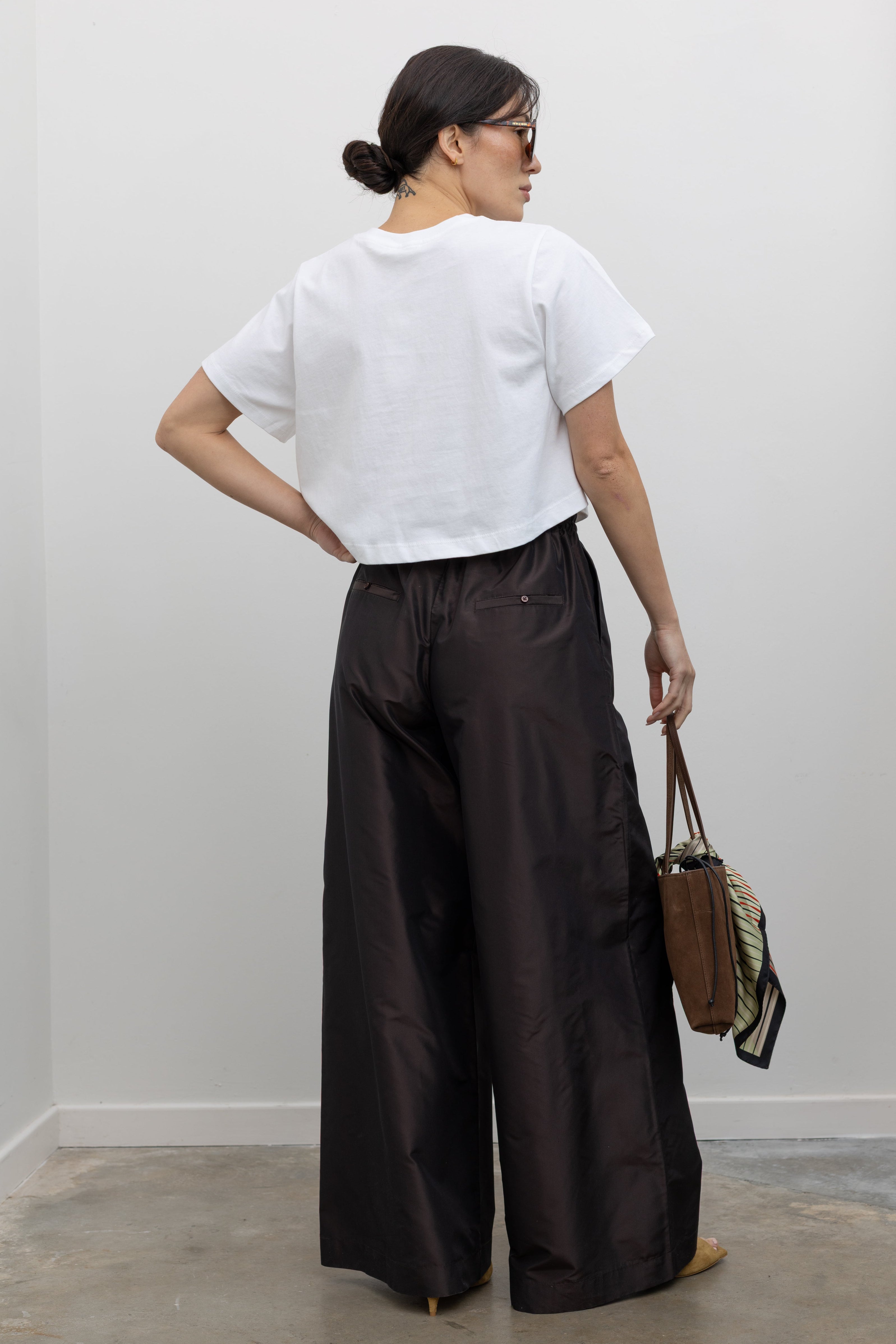 Full length back view of a woman wearing versatile and contemporary spring summer fashion in Kallmeyer Espresso Silk Taffeta Wesley Drawstring Pants featuring a hardware drawstring waist and structured wide legs at an Alas Eius store. Made in New York.