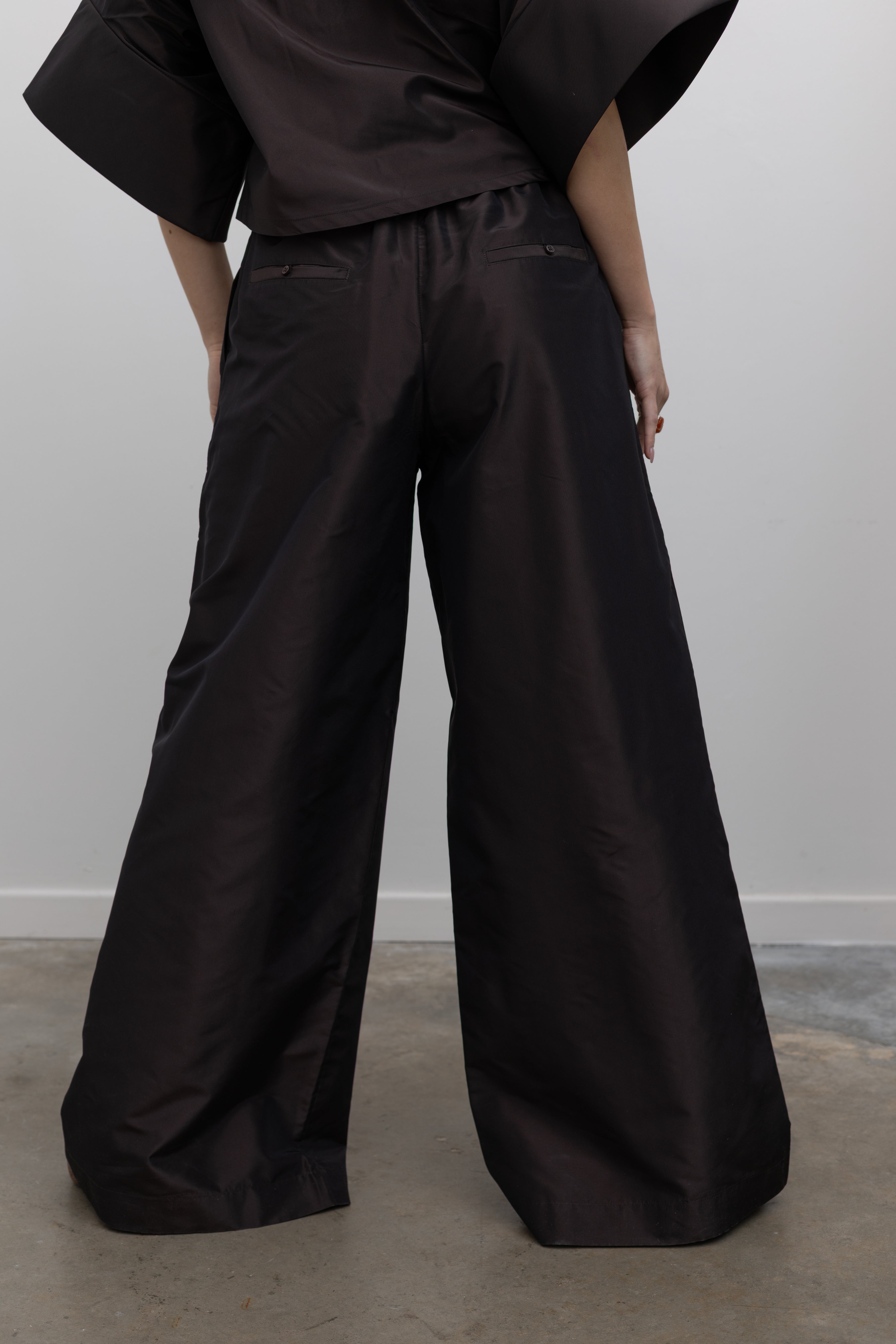 Close-up back view of a woman wearing elegant and contemporary spring fashion in Kallmeyer Espresso Silk Taffeta Wesley Drawstring Pants featuring a hardware drawstring waist and structured wide legs at an Alas Eius store. Made in New York.