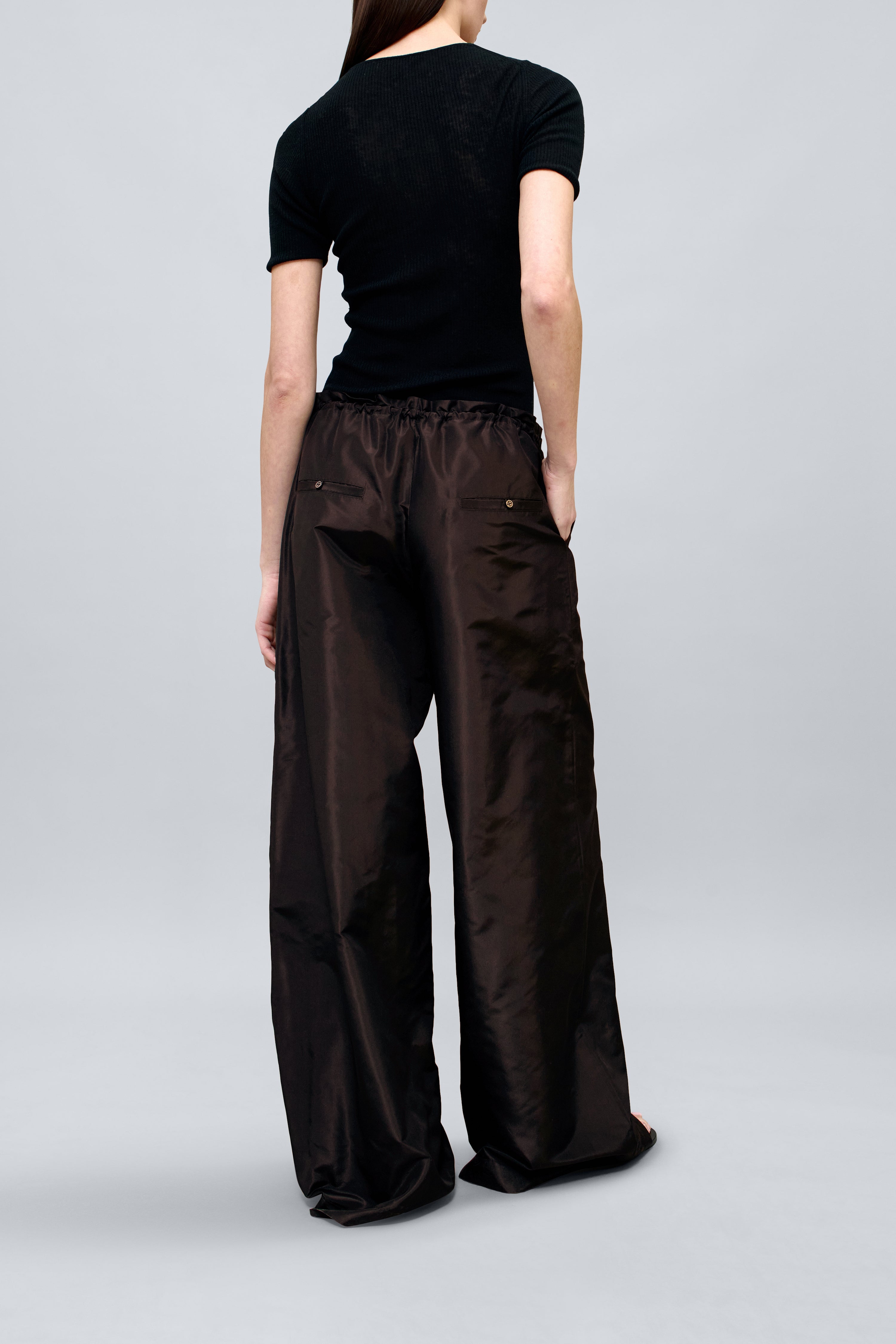 Back view of a woman wearing versatile and contemporary spring summer fashion in Kallmeyer Espresso Silk Taffeta Wesley Drawstring Pants featuring a hardware drawstring waist and structured wide legs at an Alas Eius store. Made in New York.