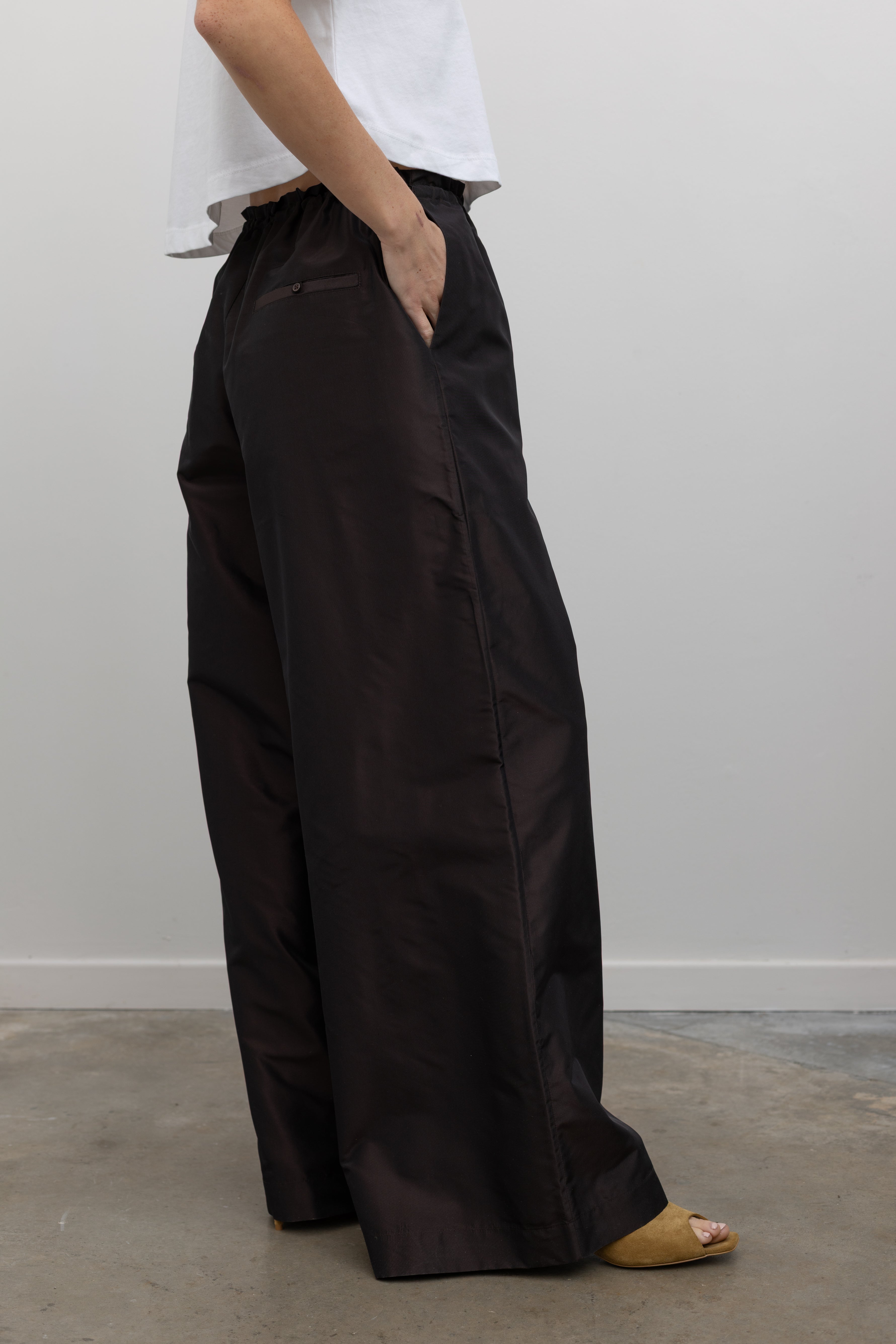 Close-up side view of a woman wearing versatile and contemporary spring summer fashion in Kallmeyer Espresso Silk Taffeta Wesley Drawstring Pants featuring a hardware drawstring waist and structured wide legs at an Alas Eius store. Made in New York.