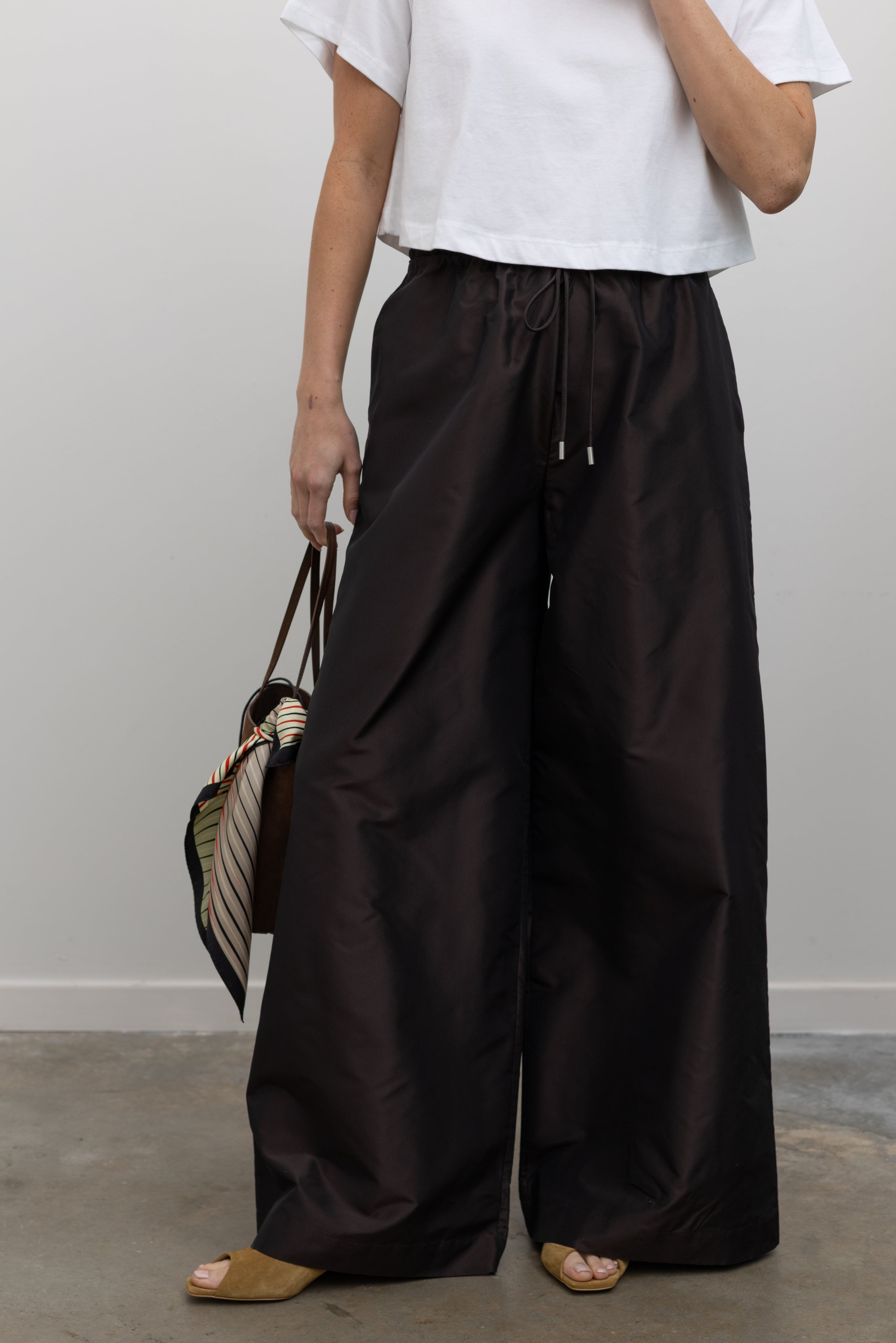 Close-up front view of a woman wearing versatile and contemporary spring summer fashion in Kallmeyer Espresso Silk Taffeta Wesley Drawstring Pants featuring a hardware drawstring waist and structured wide legs at an Alas Eius store. Made in New York.