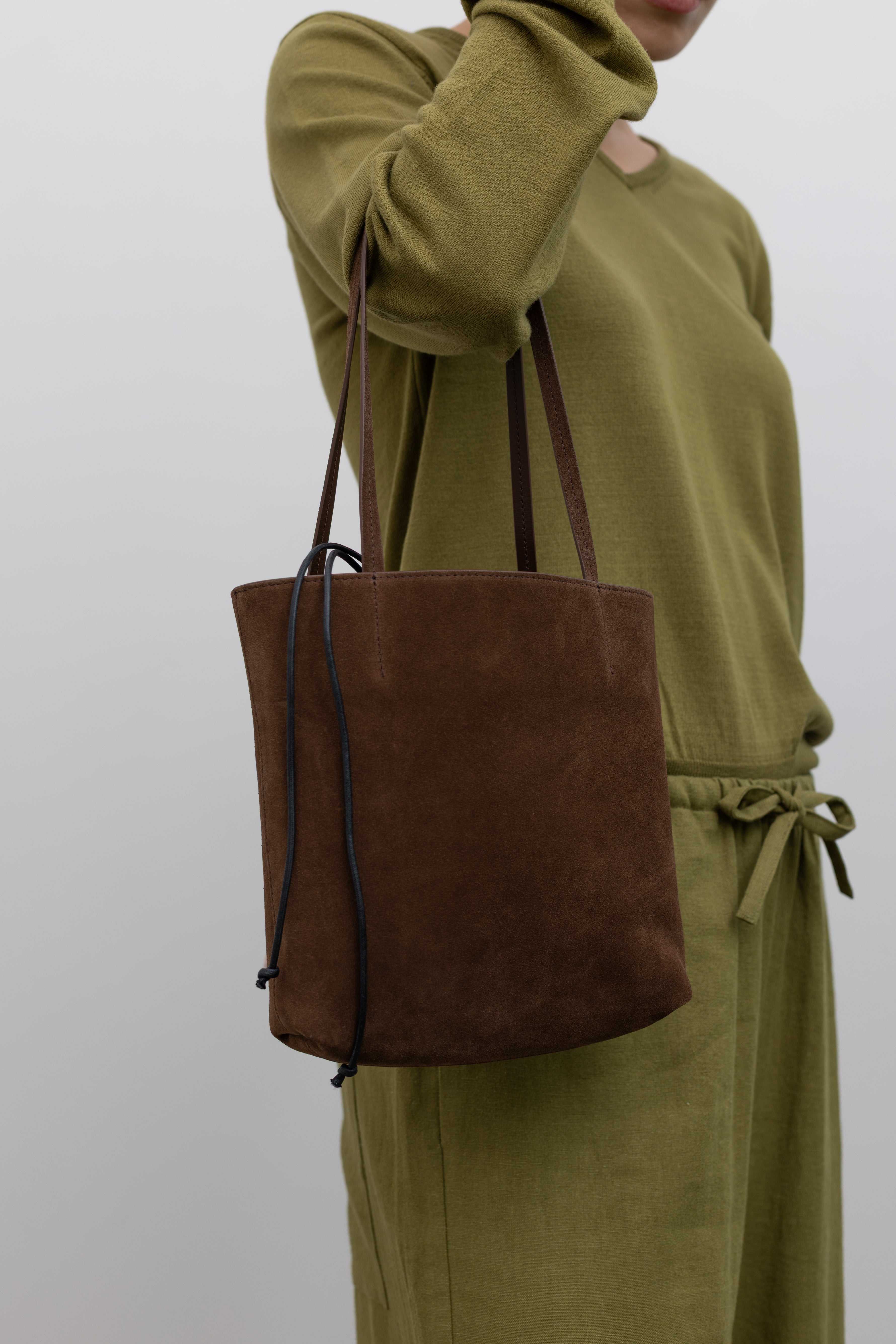 Woman carrying a sophisticated structured tote by Kallmeyer in rich brown suede, dual flat shoulder straps, and a permanent canvas drawstring pouch opening with leather toggles at Alas Eius. Made in Italy. 