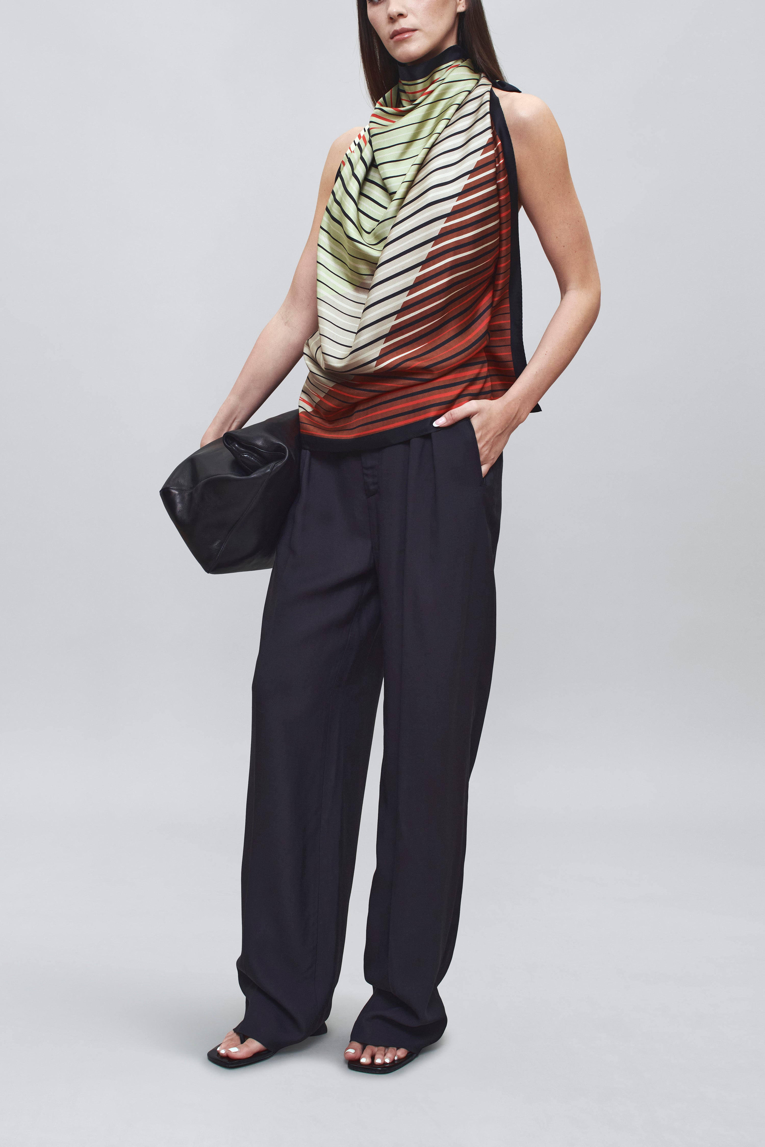 Front view of a woman wearing chic spring summer fashion in a colorful Kallmeyer Neutral Silk Twill Fluted Scarf Top featuring a gradient stripe print, soft cowl neckline, and a deep open back with fitted panel at lower back paired with navy blue trousers at an Alas Eius store.