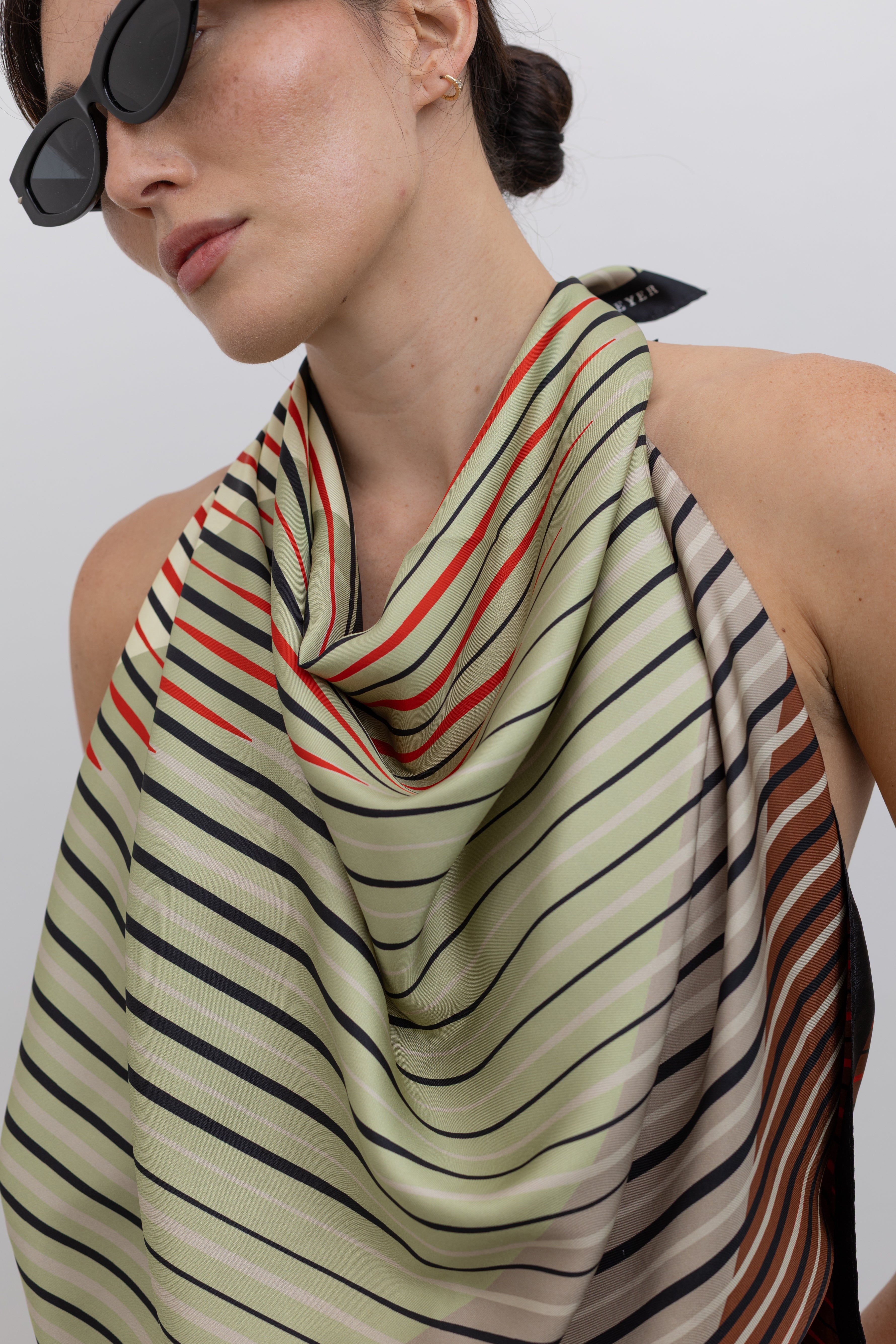 Close-up front view of a woman wearing the perfect summer vacation wardrobe staple in a colorful Kallmeyer Neutral Silk Twill Fluted Scarf Top featuring a gradient stripe print, soft cowl neckline, and a deep open back at an Alas Eius store.