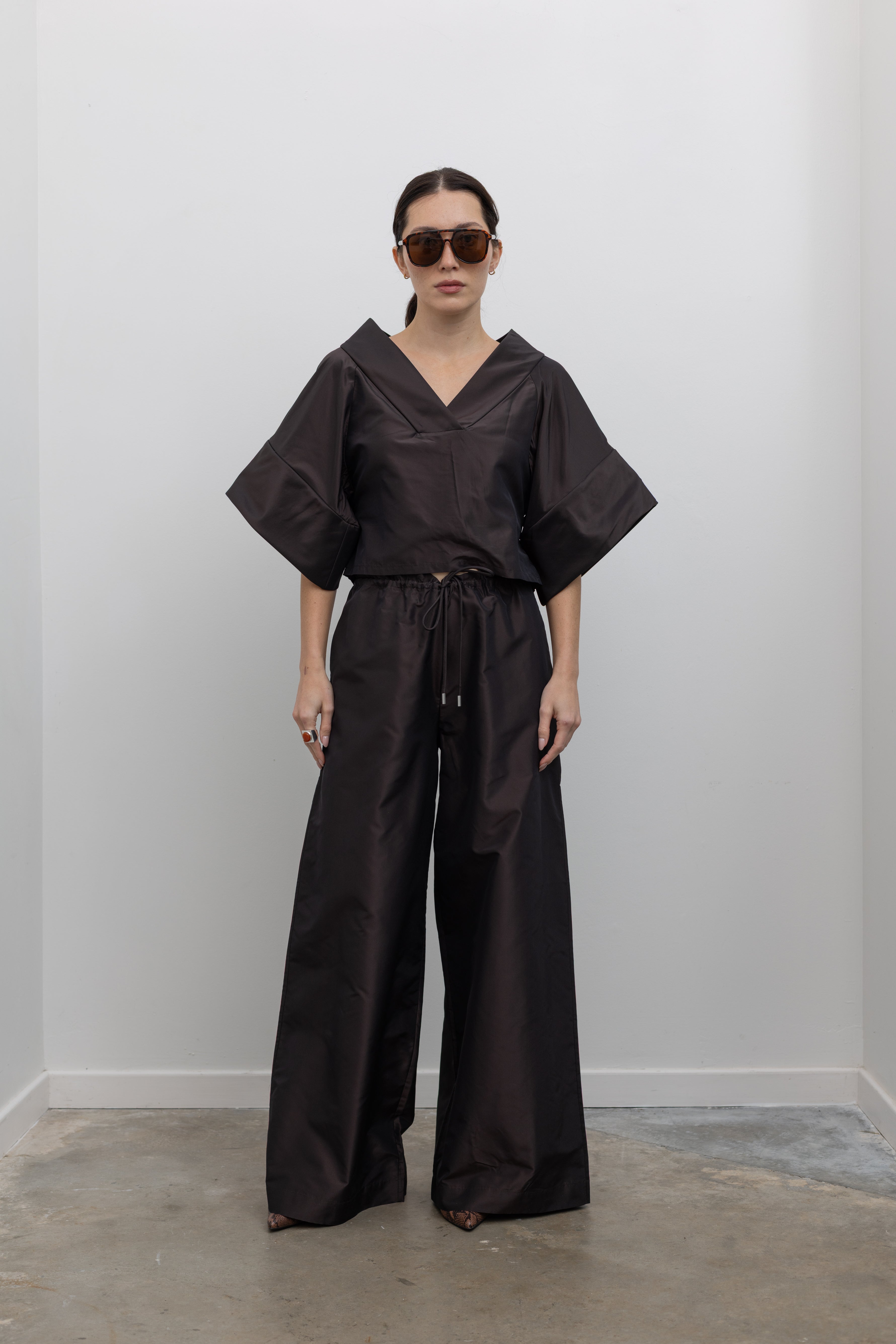 Full length front view of a woman wearing chic and contemporary fashion in a Kallmeyer Espresso Silk Taffeta Winslow Top featuring a dramatic structured v-neckline and oversized cuffed tunic sleeves; paired with Espresso Silk Taffeta Wesley Drawstring Pant featuring a hardware drawstring waist and structured wide legs at an Alas Eius store. Made in New York.