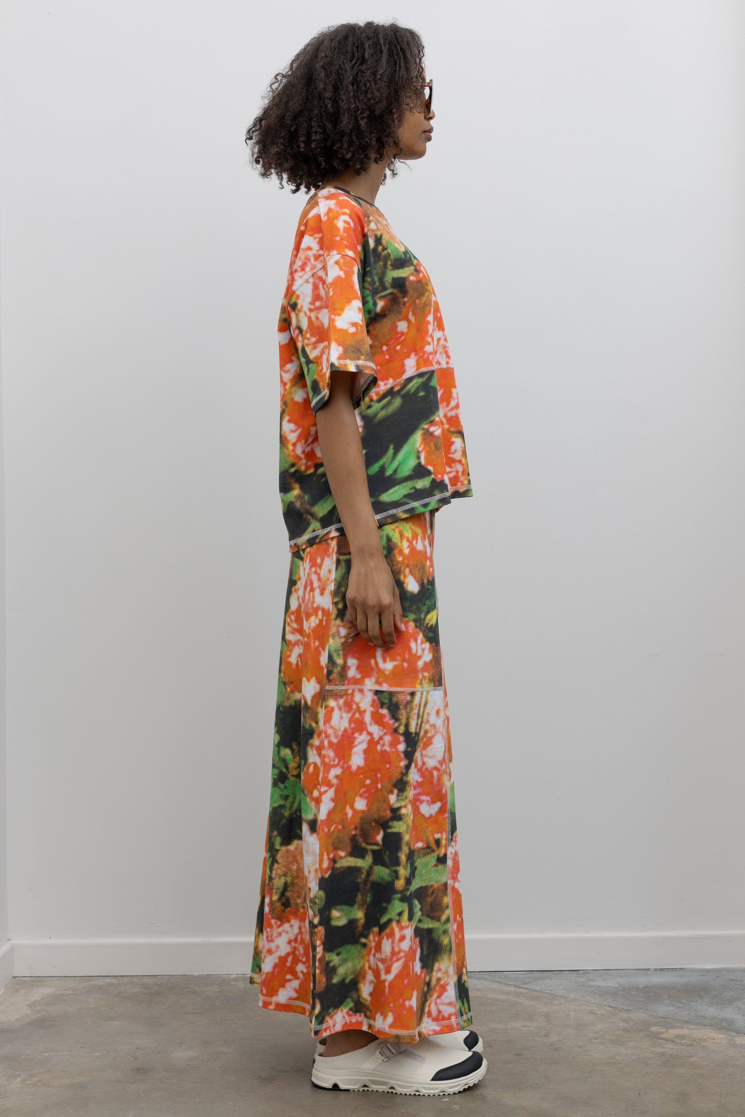 Full length front view of a woman wearing the perfect travel set in a vibrant and abstract print Kowtow Flower Book Montage Tee featuring a boxy shape, drop shoulders, and patchwork panelling throughout that is highlighted with contrast white top stitching paired with its matching Flower Book Montage maxi skirt at an Alas Eius store. 