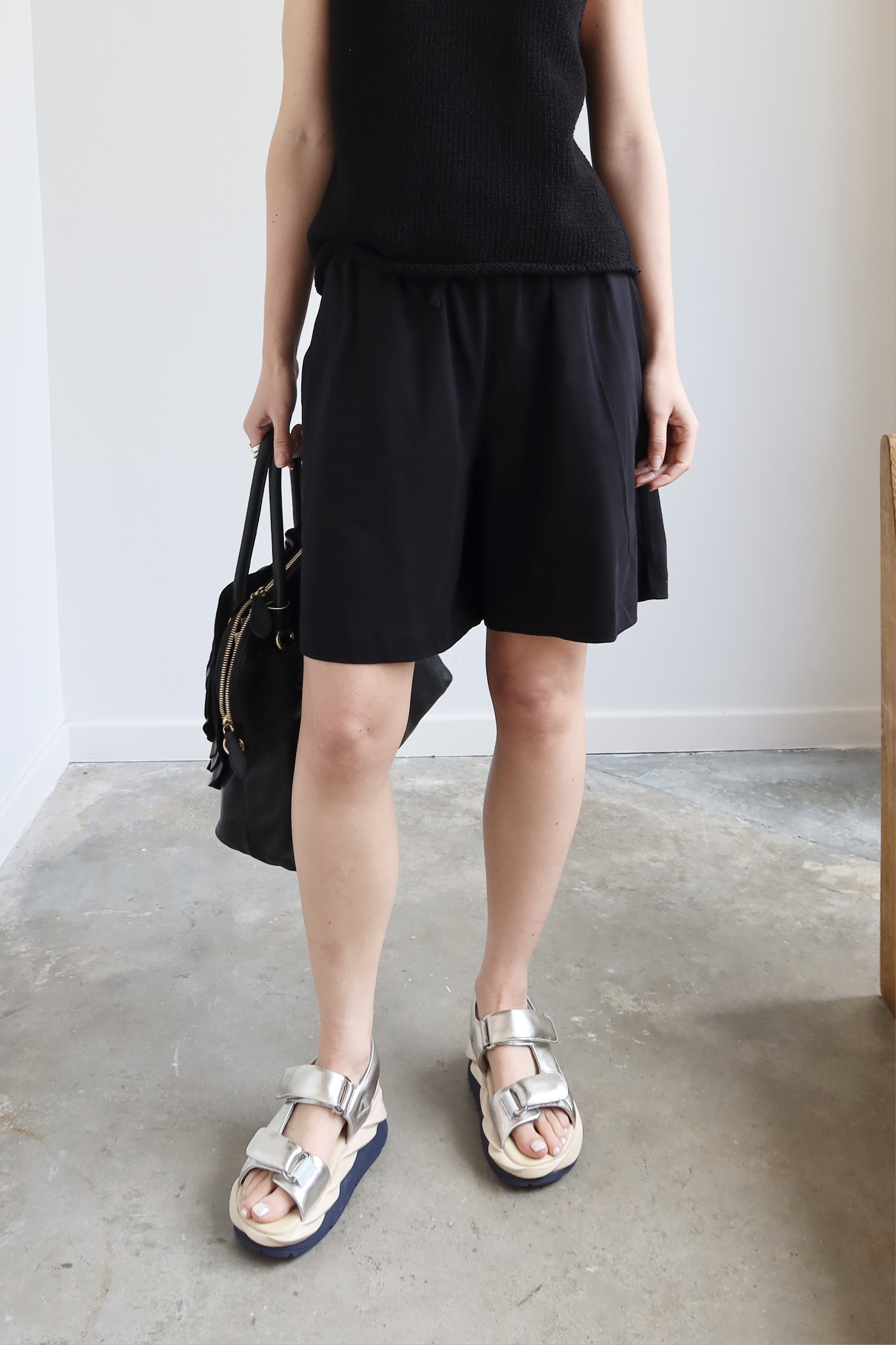 Close-up front view of a woman standing wearing spring fashion in a sophisticated pair of Kowtow black organic fairtrade cotton drawstring shorts with a black sleeveless top and sandals at Alas Eius. 