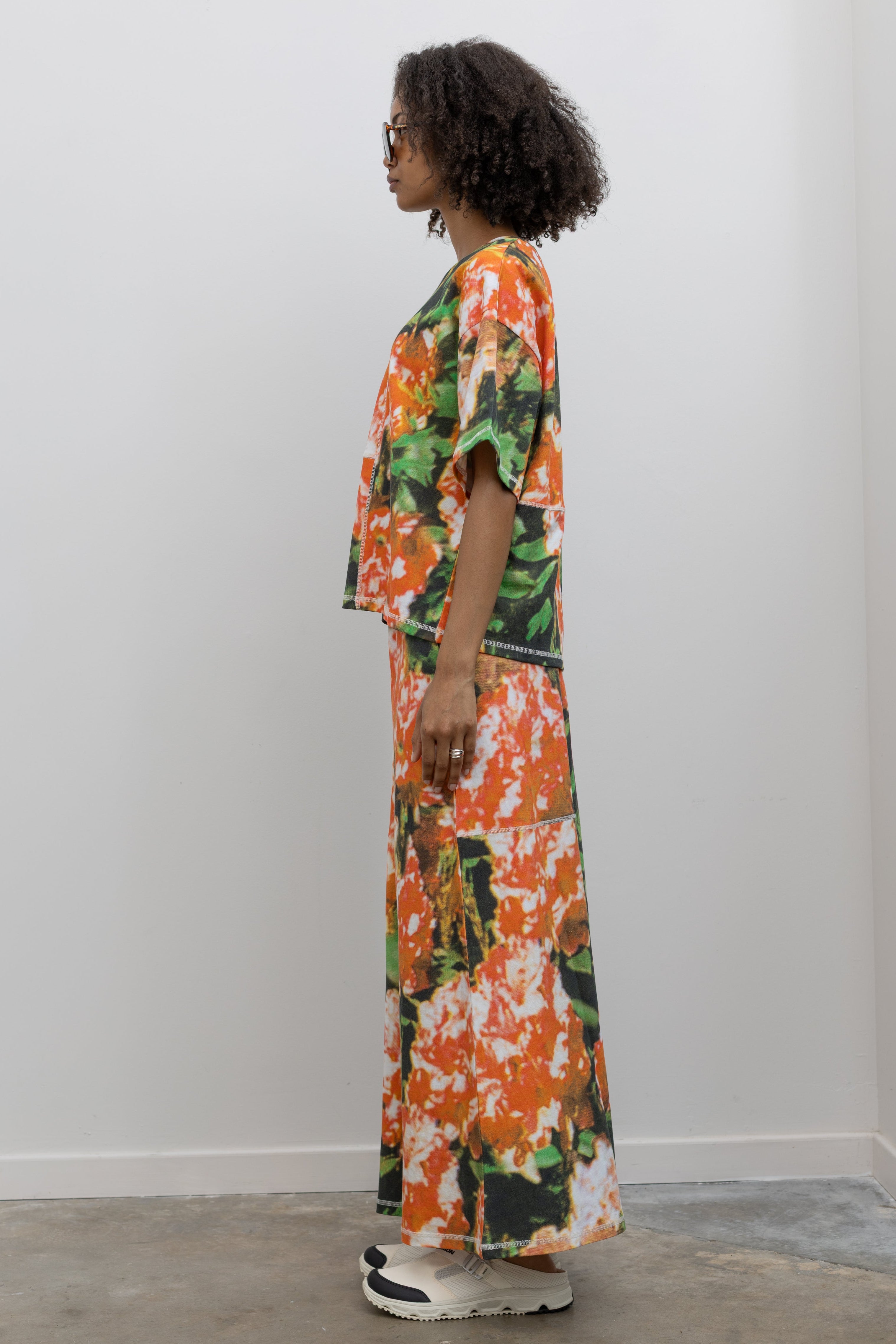 Full length side view of a woman wearing the perfect travel set in a vibrant and abstract print Kowtow Flower Book Montage Tee featuring a boxy shape, drop shoulders, and patchwork panelling throughout that is highlighted with contrast white top stitching paired with its matching Flower Book Montage maxi skirt at an Alas Eius store. 