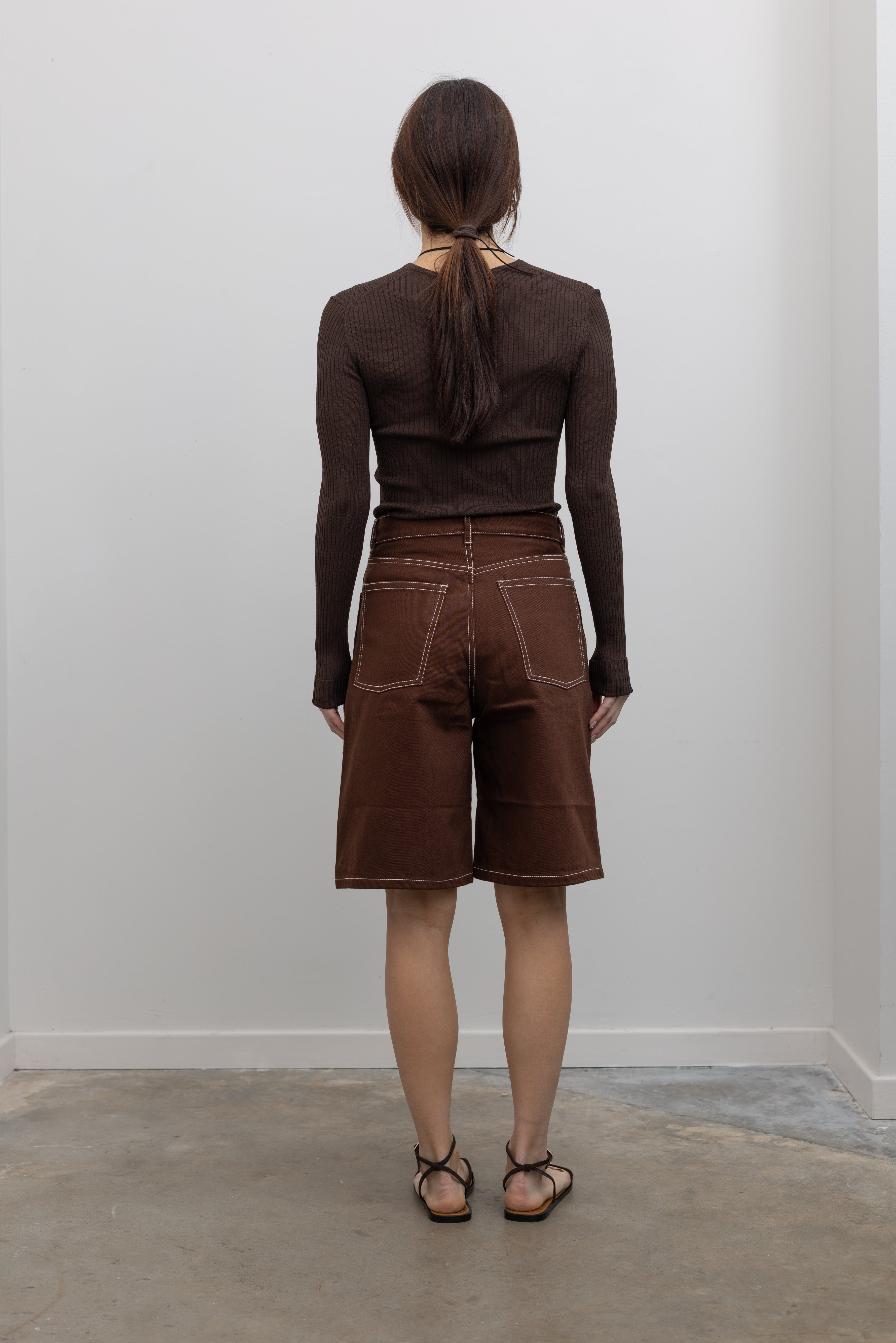 Back view of Woman wearing Spring fashion in a pair of Kowtow Framework Brown relaxed, straight fit Denim Shorts with contrast white topstitching and brown long-sleeved ribbed top with a shell necklace and sunglasses at Alas Eius