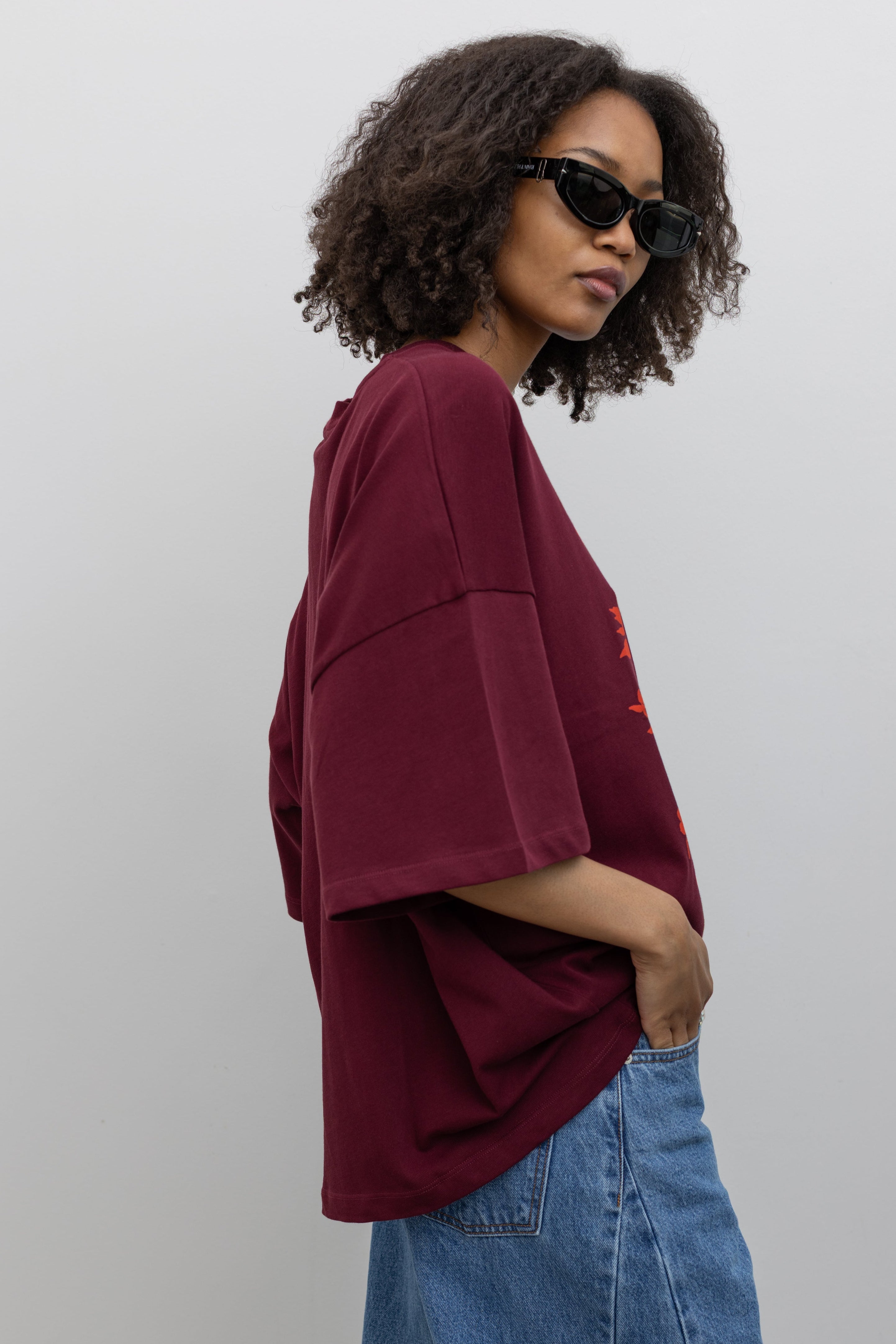 Side view of a woman posing wearing an elevated essential in a Kowtow Maroon Stencil Tee featuring a screen-printed pressed foliage motif with short sleeves, an oversized fit, and a ribbed crew neck paired with light wash relaxed fit denim jeans at an Alas Eius store. 