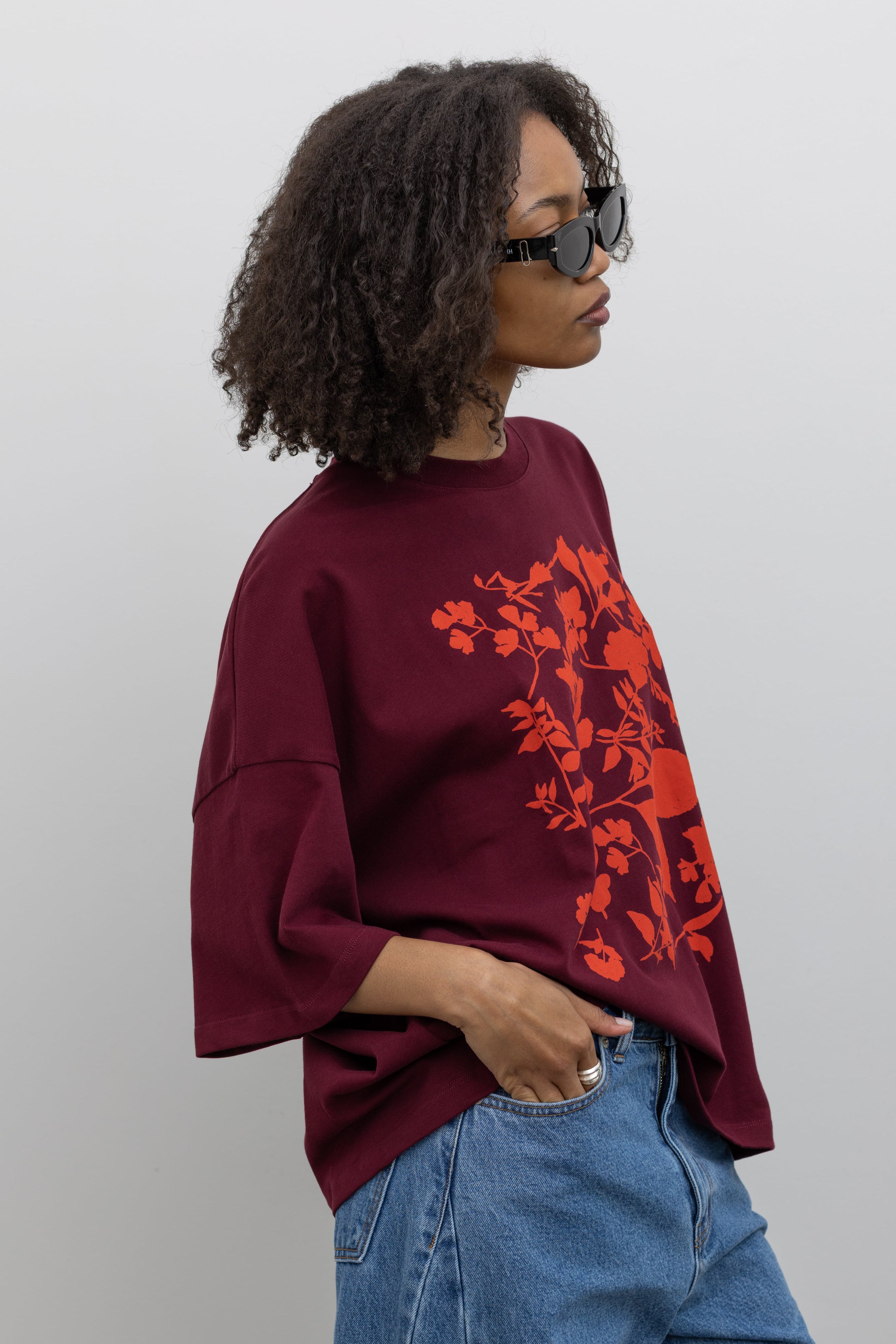 3/4 view of a woman posing wearing a summer wardrobe essential in a Kowtow Maroon Stencil Tee featuring a screen-printed pressed foliage motif with short sleeves, an oversized fit, and a ribbed crew neck paired with light wash relaxed fit denim jeans at an Alas Eius store. 