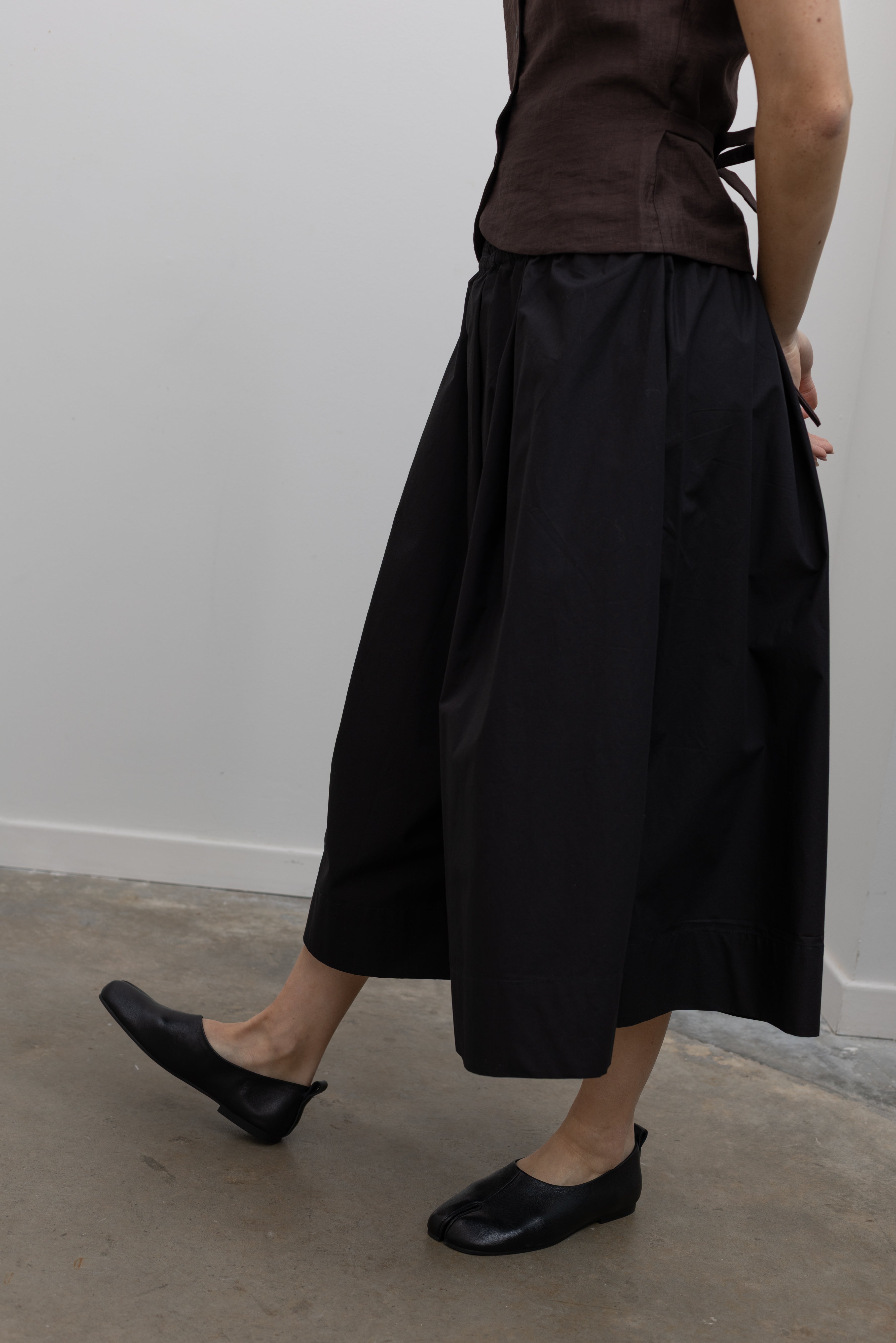 Detailed product side view of Woman wearing Spring fashion in a brown linen buttoned vest with flattering Kowtow Black Organic Cotton Elastic Waistband Culottes with black leather flat shoes at Alas Eius