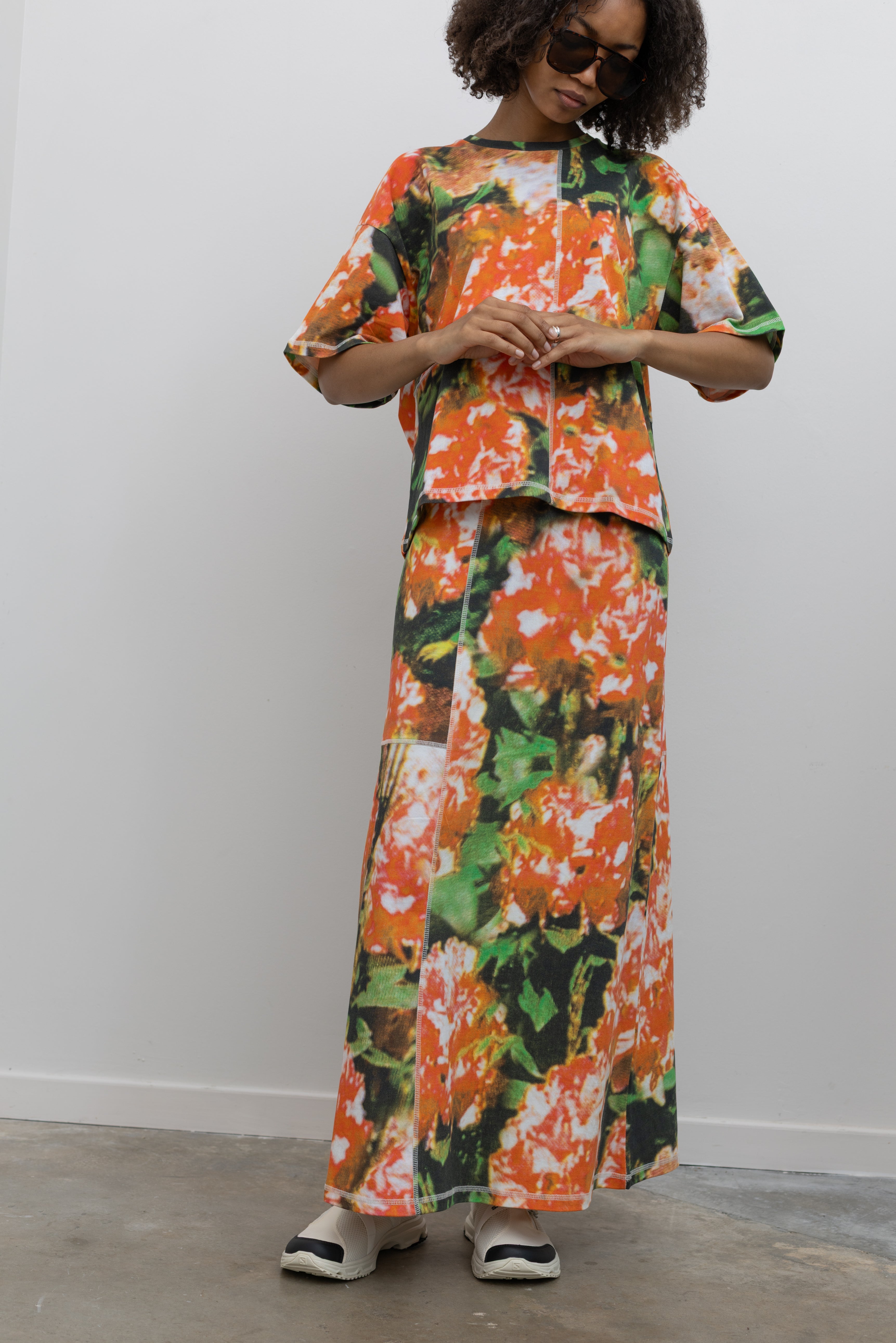 Full length front view of a woman wearing an elevated set in a vibrant and abstract print Kowtow Flower Book Montage Tee featuring a boxy shape, drop shoulders, and patchwork panelling throughout that is highlighted with contrast white top stitching paired with the Flower Book Montage maxi skirt at an Alas Eius store. 