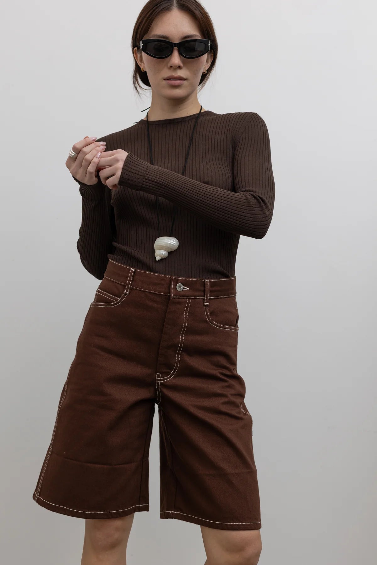 Woman wearing Spring fashion in a pair of Kowtow Framework Brown relaxed, straight fit Organic Cotton Denim Shorts with contrast white topstitching and brown long-sleeved ribbed top with a shell necklace and sunglasses Alas Eius