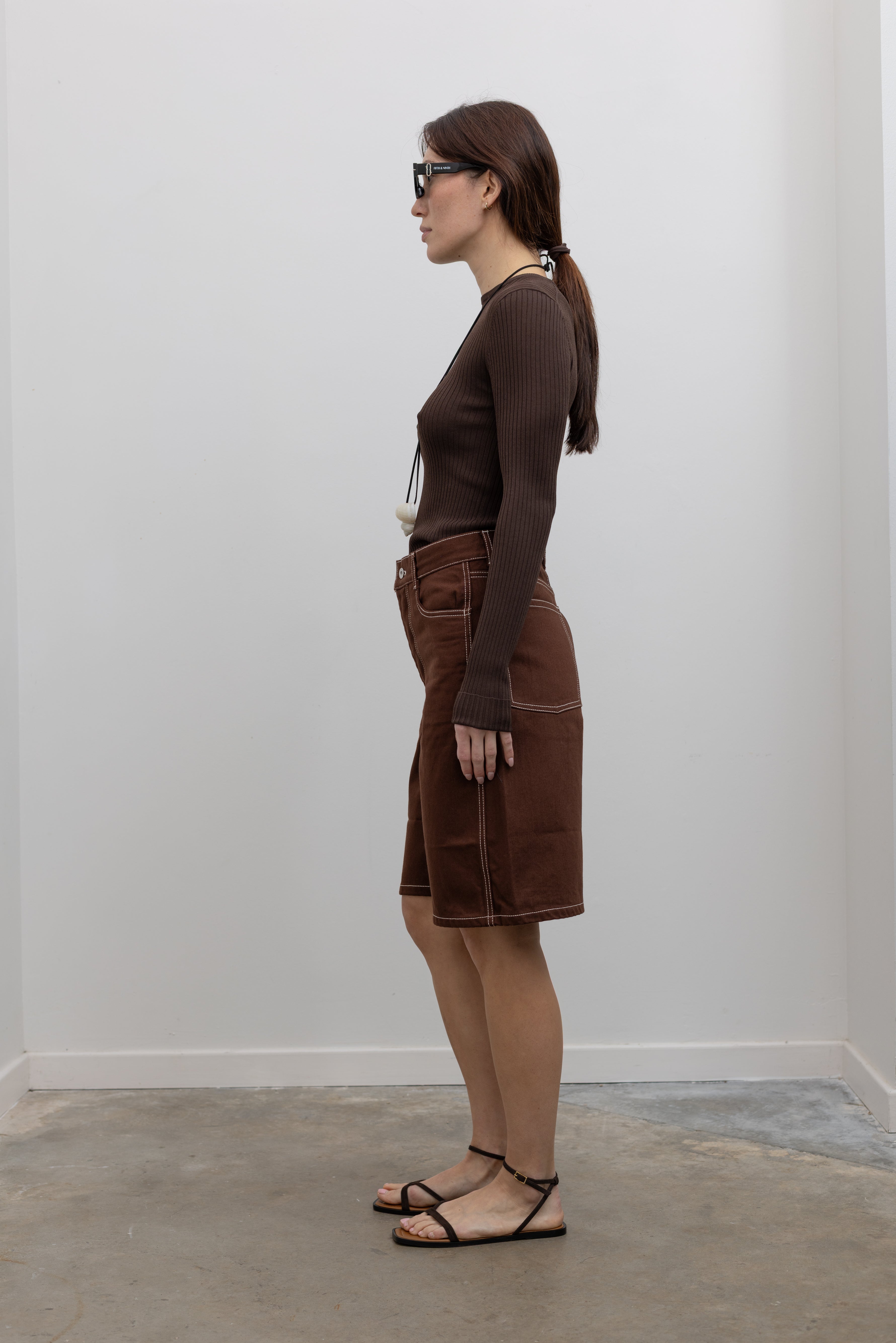 Side view of Woman wearing Spring fashion in a pair of Kowtow Framework Brown relaxed, straight fit Organic Cotton Denim Shorts with contrast white topstitching and brown long-sleeved ribbed top with a shell necklace and sunglasses at Alas Eius