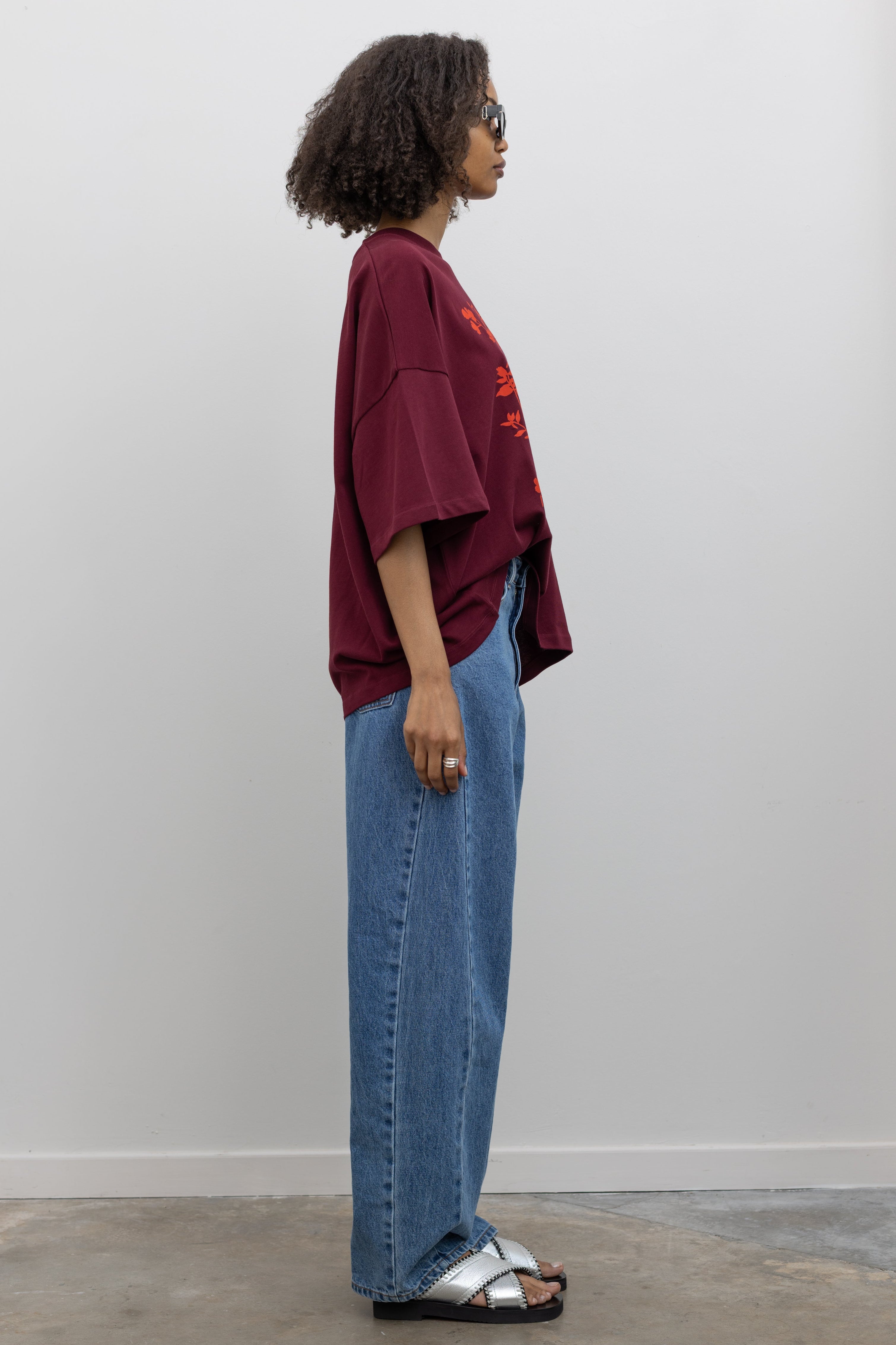 Full length side view of a woman posing wearing an elevated essential in a Kowtow Maroon Stencil Tee featuring a screen-printed pressed foliage motif with short sleeves, an oversized fit, and a ribbed crew neck paired with light wash relaxed fit denim jeans at an Alas Eius store. 