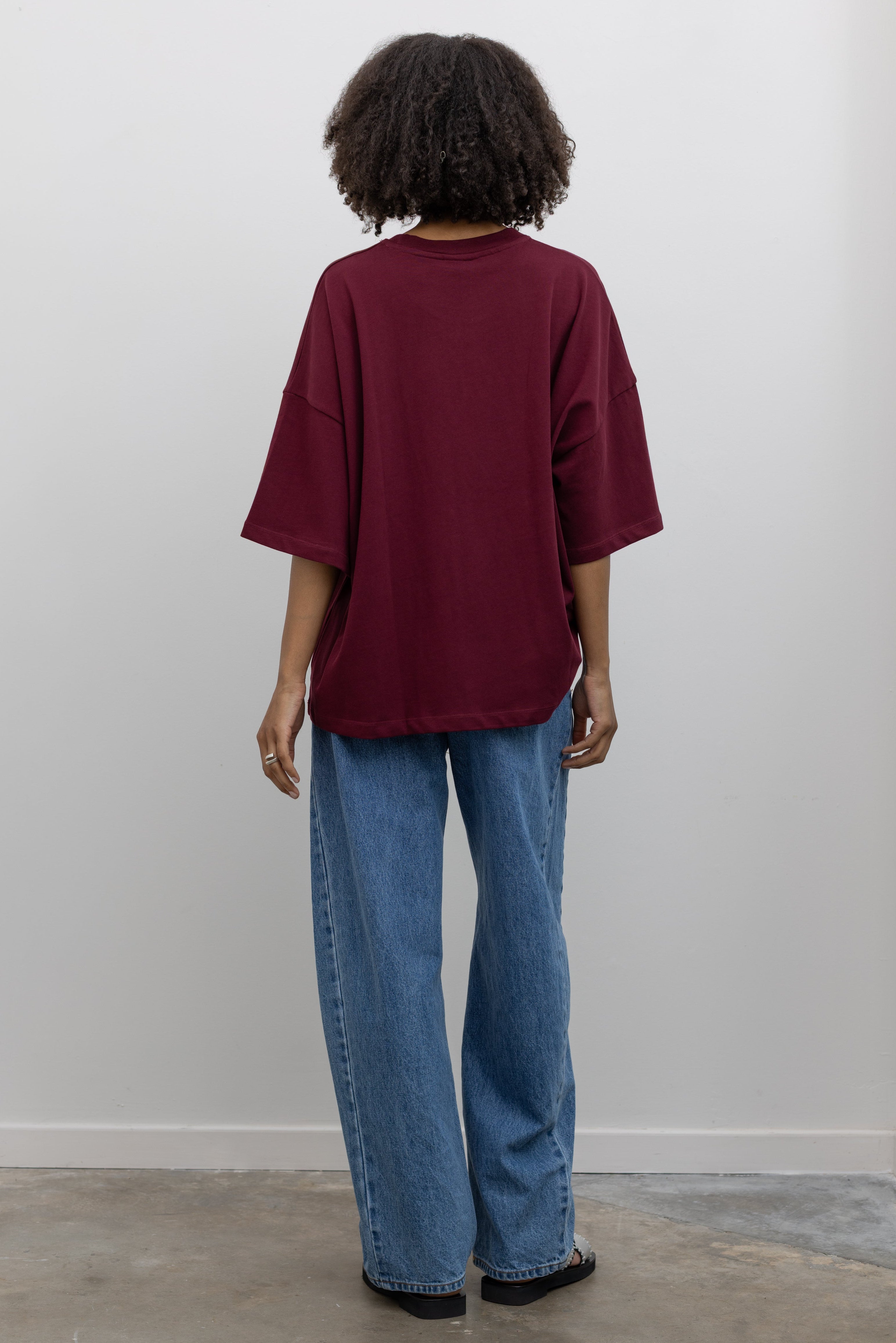 Full length back view of a woman posing wearing an elevated essential in a Kowtow Maroon Stencil Tee featuring a screen-printed pressed foliage motif with short sleeves, an oversized fit, and a ribbed crew neck paired with light wash relaxed fit denim jeans at an Alas Eius store. 