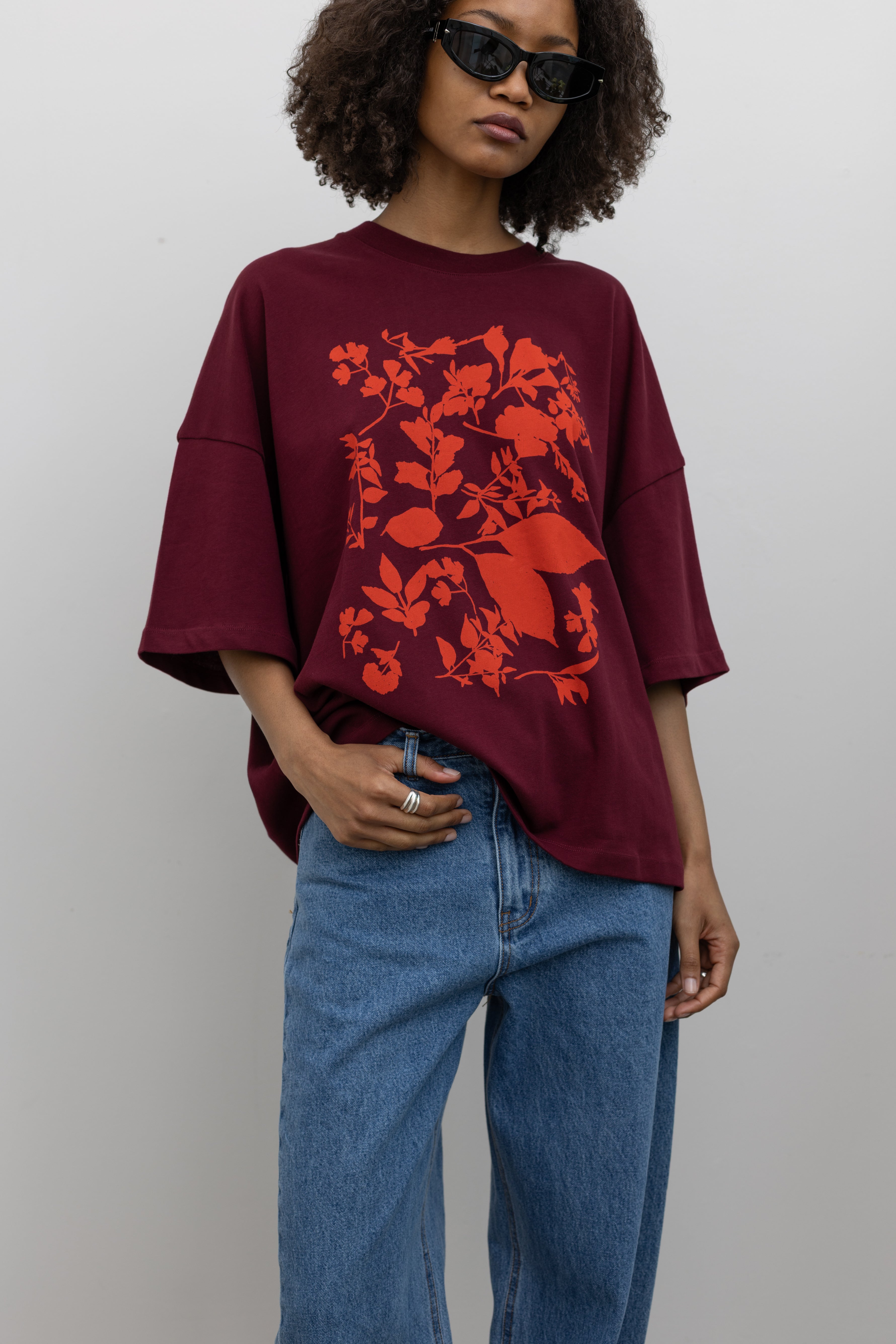 Front view of a woman posing wearing an elevated essential in a Kowtow Maroon Stencil Tee featuring a screen-printed pressed foliage motif with short sleeves, an oversized fit, and a ribbed crew neck paired with light wash relaxed fit denim jeans at an Alas Eius store. 