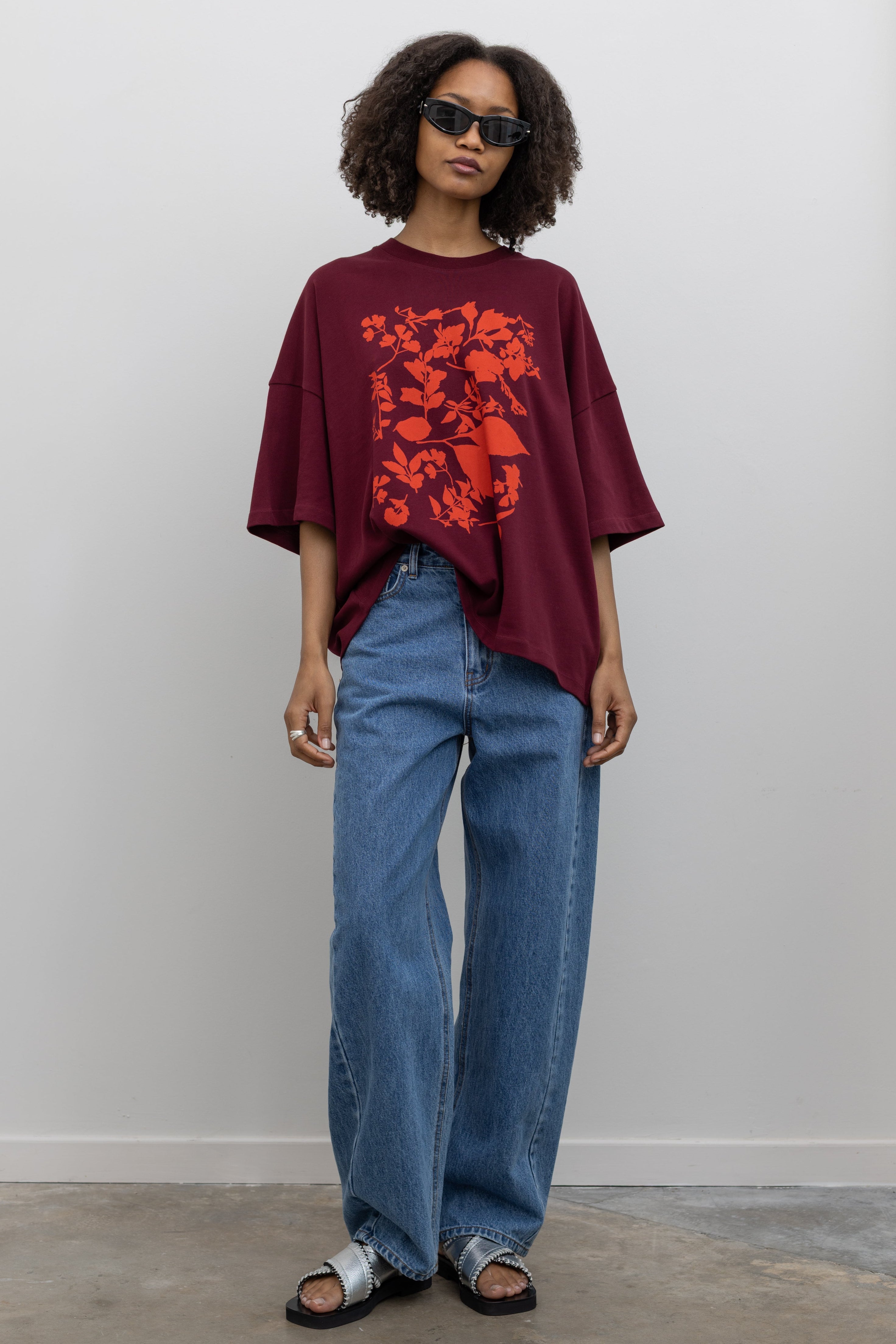 Full length front view of a woman posing wearing an elevated essential in a Kowtow Maroon Stencil Tee featuring a screen-printed pressed foliage motif with short sleeves, an oversized fit, and a ribbed crew neck paired with light wash relaxed fit denim jeans at an Alas Eius store. 