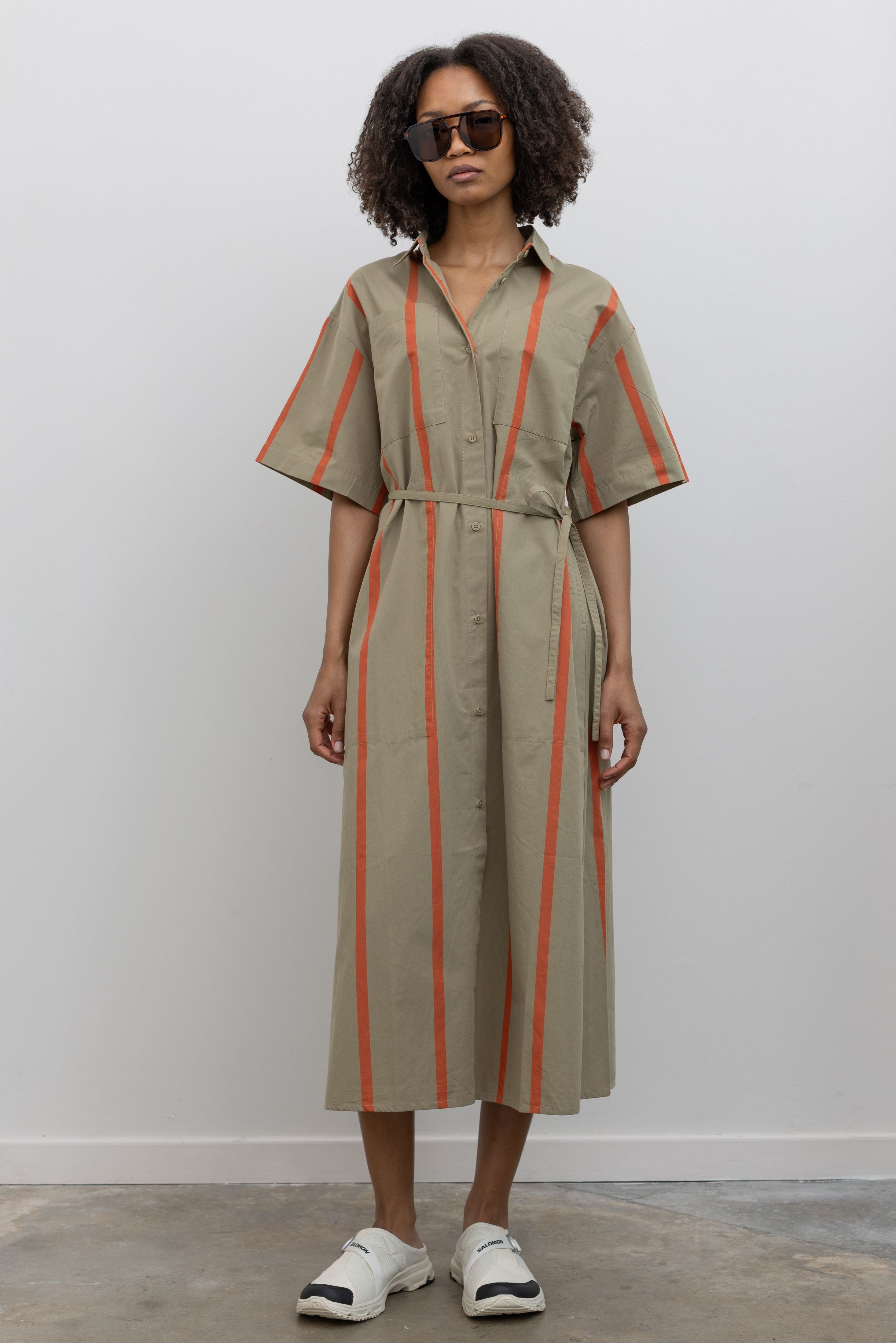 Full length front view of a woman standing wearing modern minimalist spring fashion in a Kowtow Nasturtium Stripe Oversized Balance Dress featuring pockets, a classic collar, nut buttons down the front, and a tie belt paired with Salomon slip on sneakers at Alas Eius. 