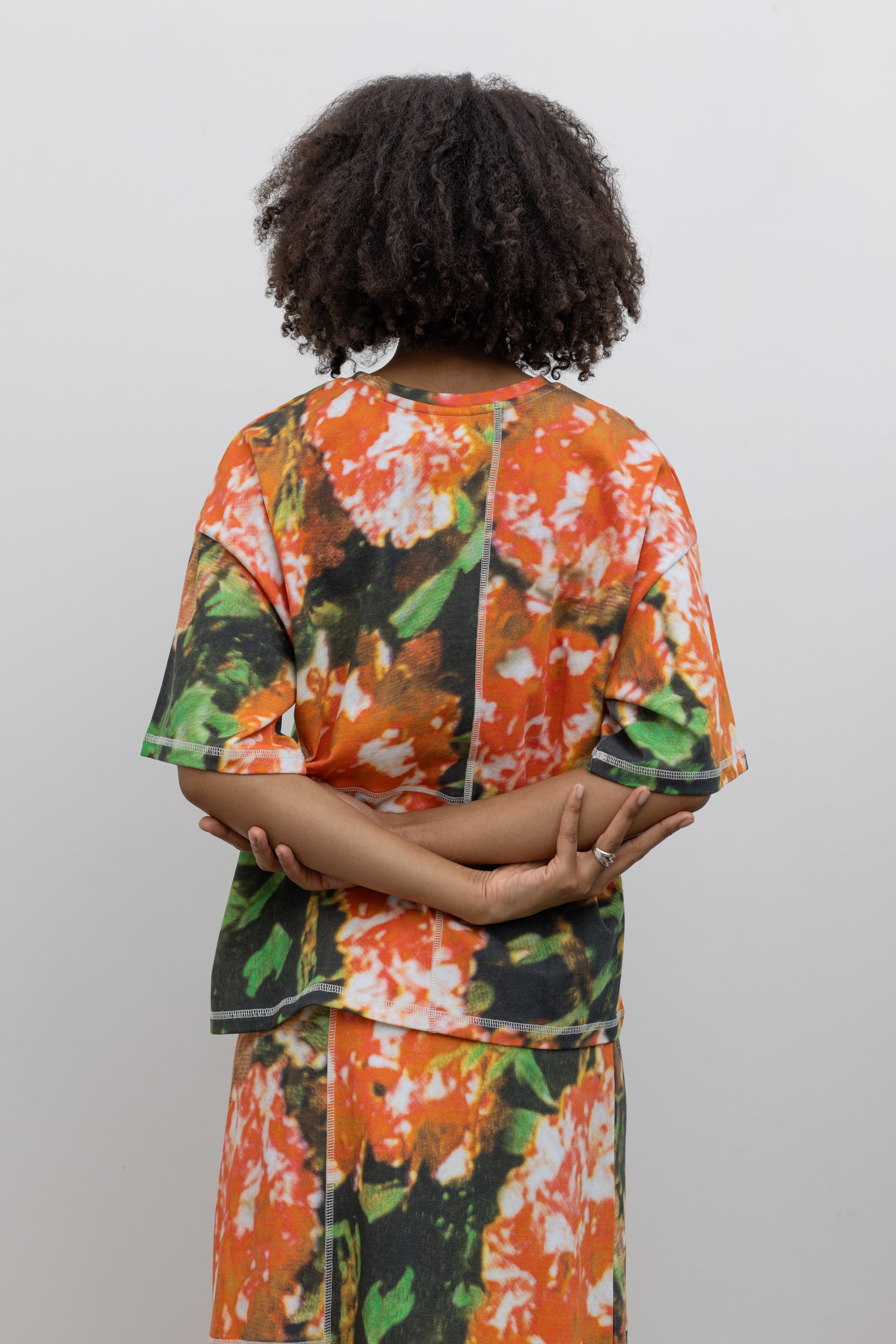 Back view of a woman wearing the perfect travel set in a vibrant and abstract print Kowtow Flower Book Montage Tee featuring a boxy shape, drop shoulders, and patchwork panelling throughout that is highlighted with contrast white top stitching paired with its matching Flower Book Montage maxi skirt at an Alas Eius store. 
