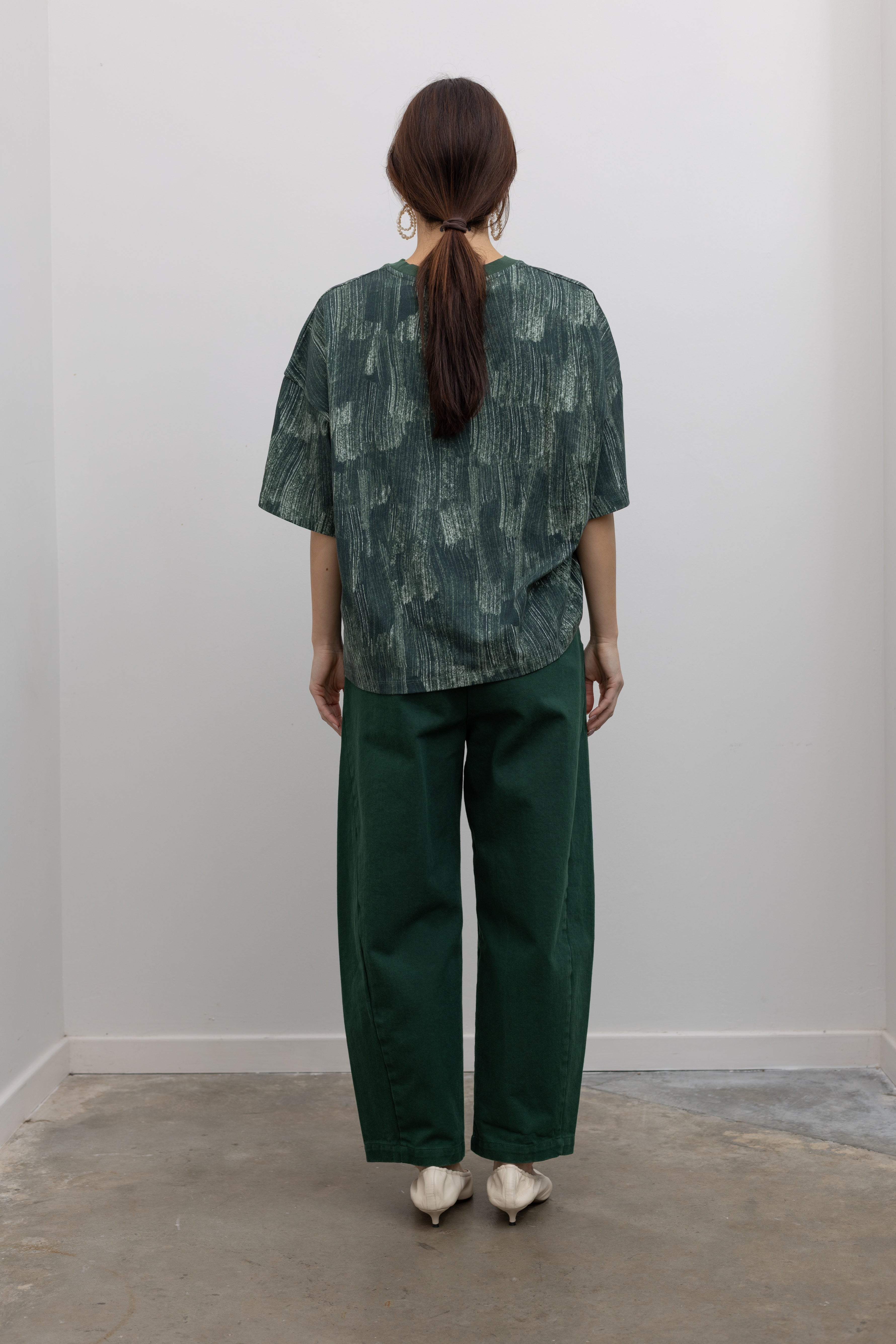Full back view of a woman posing with sunglasses wearing spring fashion in a Kowtow green oversized short sleeve t-shirt with an all over white brushstroke print paired with pine colored denim jeans at Alas Eius.